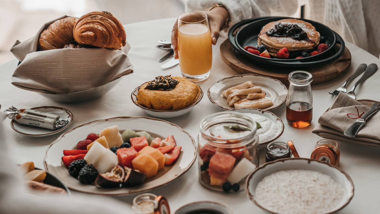 A table set with breakfast for two including plates of fresh fruit, oatmeal, pancakes, small pastries, a basket of croissants, juice and coffee
