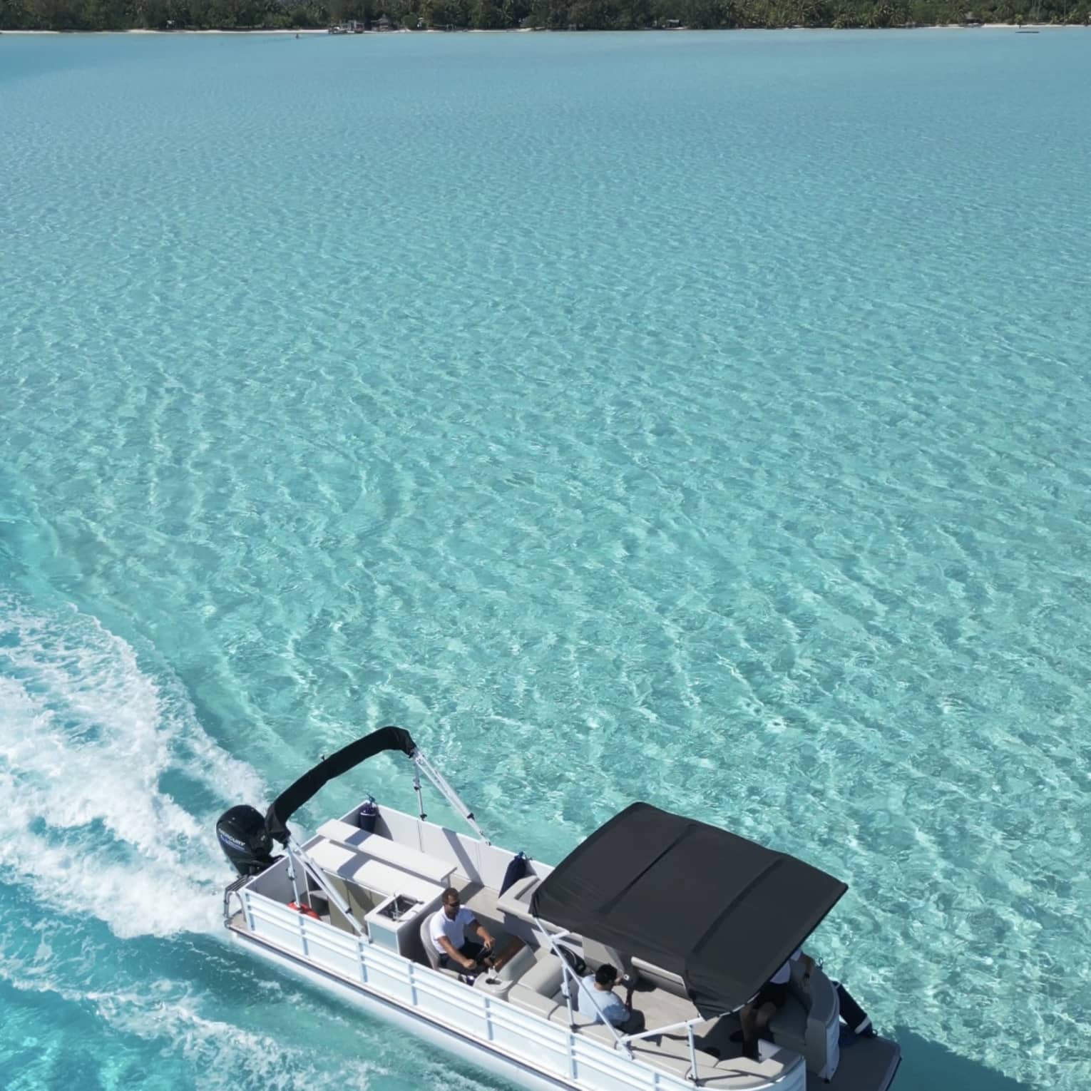 Aerial view of a pontoon boat in pale turquoise waters; guests are relaxing beneath a small canopy at the bow.