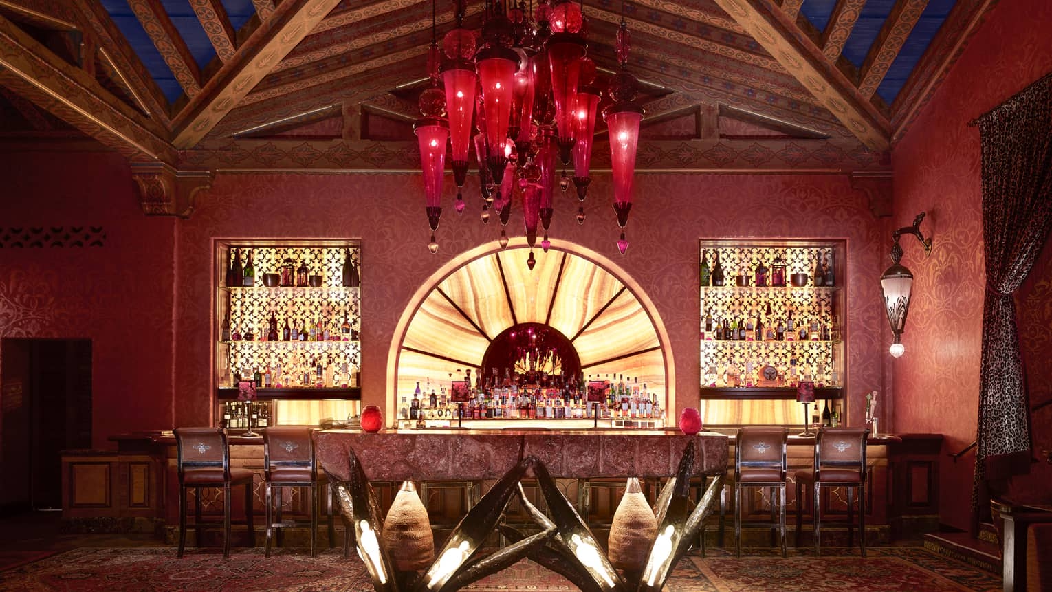 Indoor bar with illuminated shelves holding bottles, red hanging light fixture, geometric ceiling