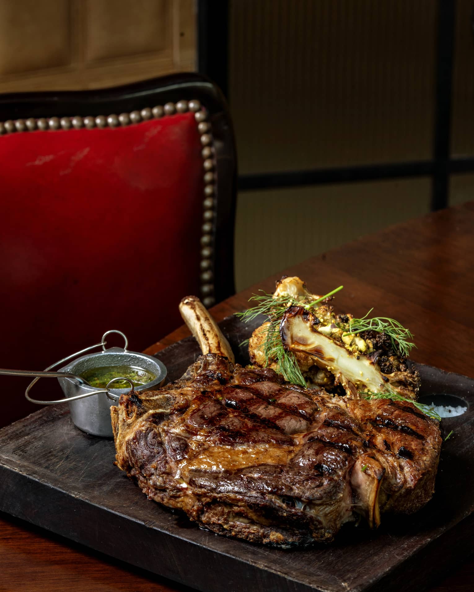 A thick, succulent cowboy ribeye steak rests on a wooden serving board, flanked by grilled vegetables and chimichurri sauce.