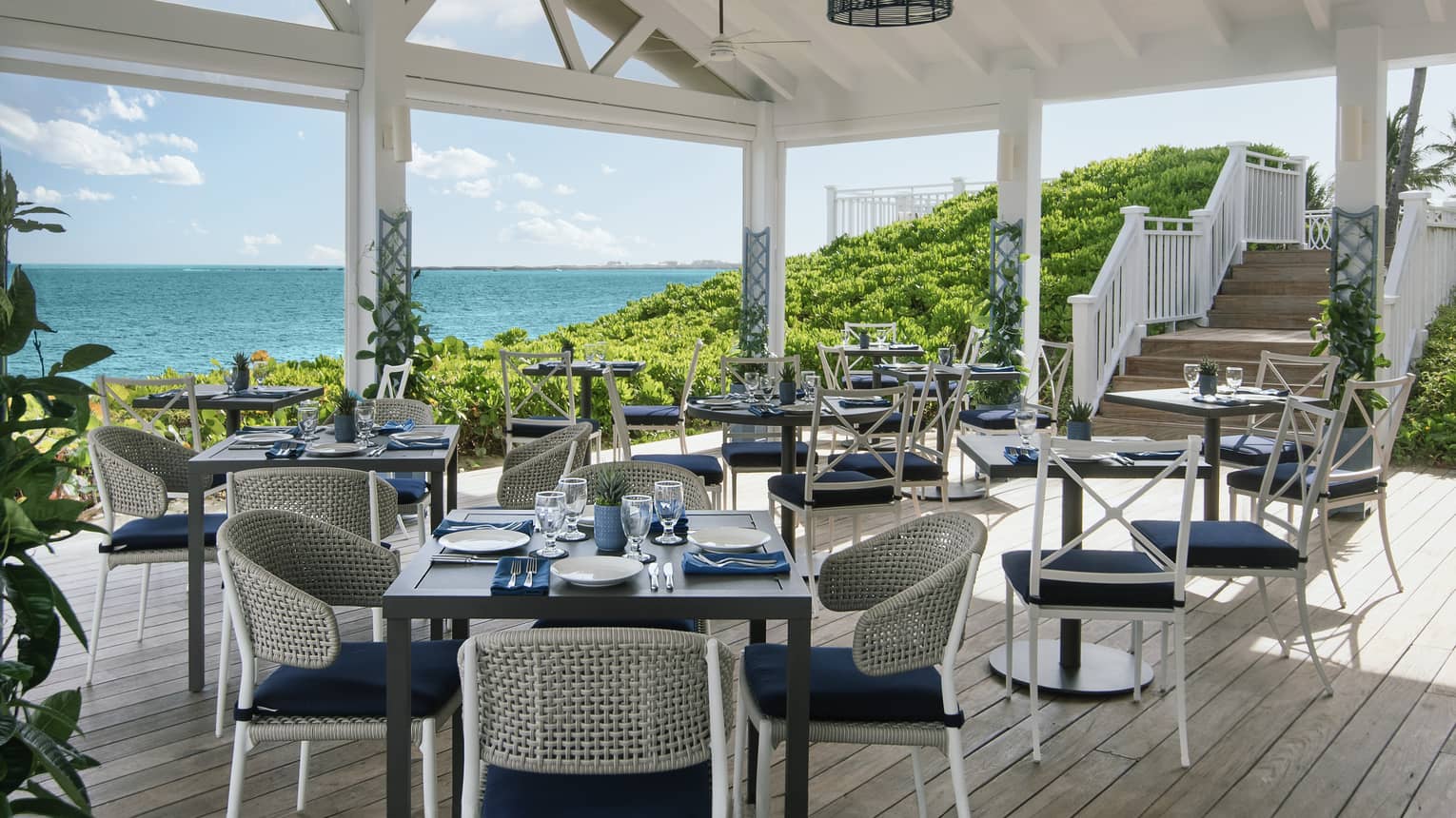 Covered restaurant patio with vasious square tables each set with four blue-and-white chairs with a view of the ocean in the background