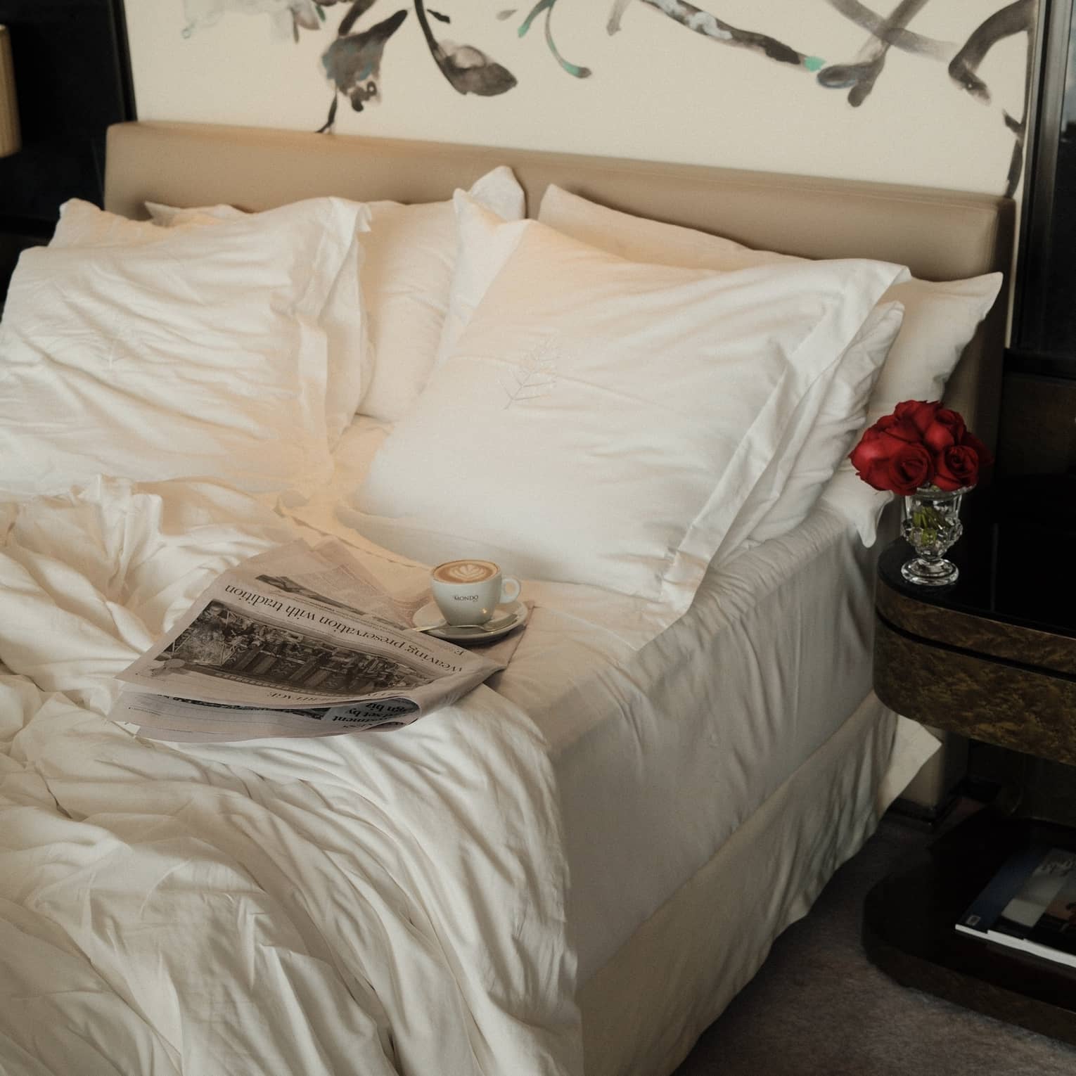 A newspaper and cup of coffee on a saucer sit atop an unmade bed with crisp, white sheets, with a pair of white slippers on the floor beneath it