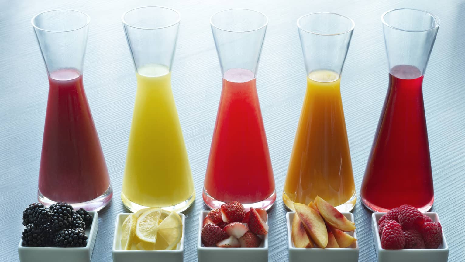 Five glass juice jugs aligned with five small trays of fruit