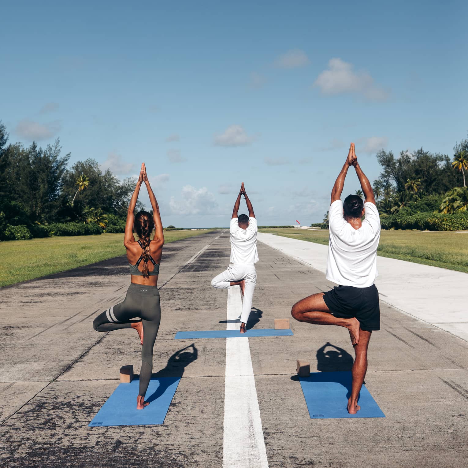 Rear view of three people practicing yoga in tree pose on an airport runway framed by lush greenery, under a clear blue sky.