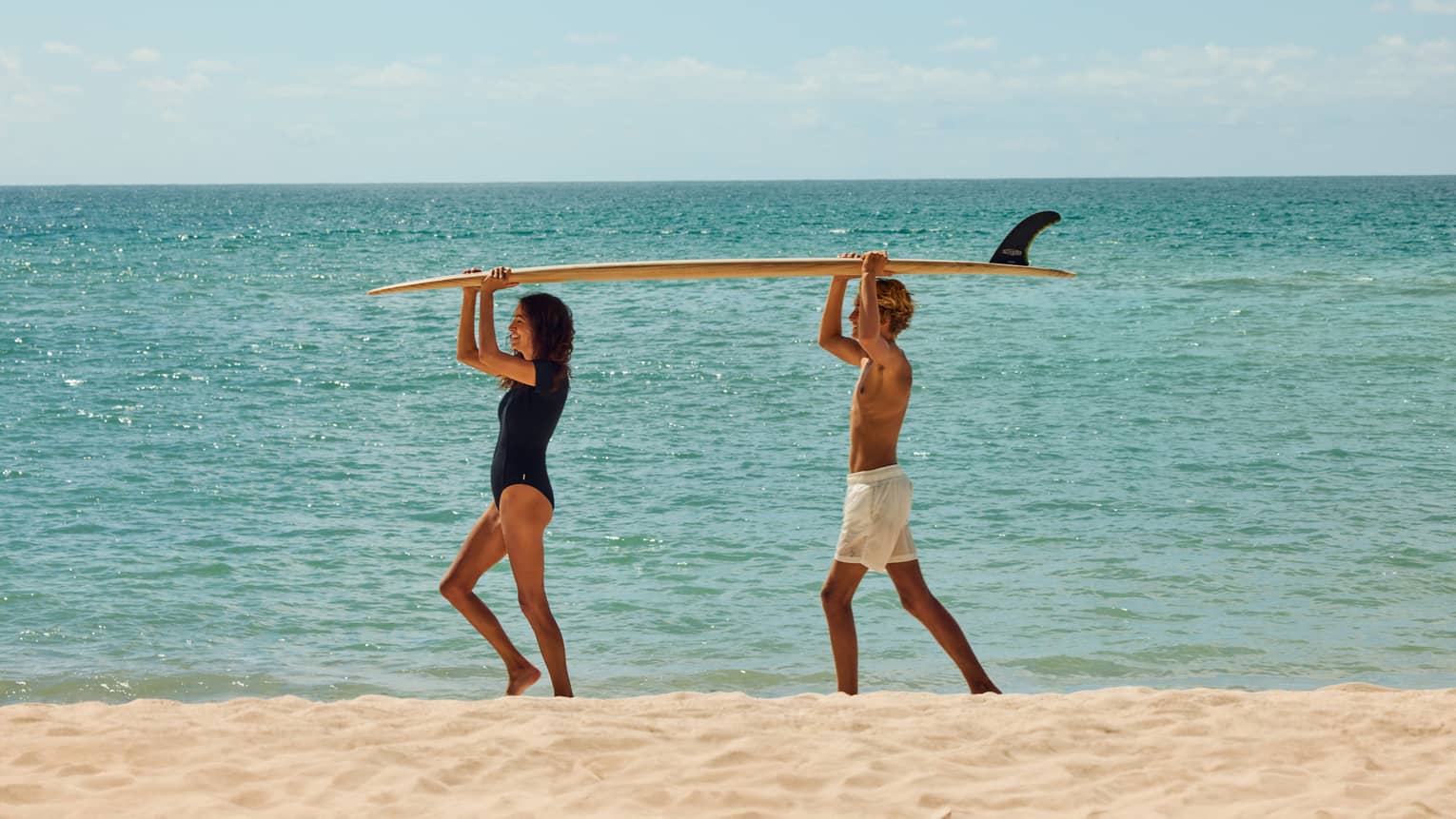 Two kids walk on beach with a surfboard on their head
