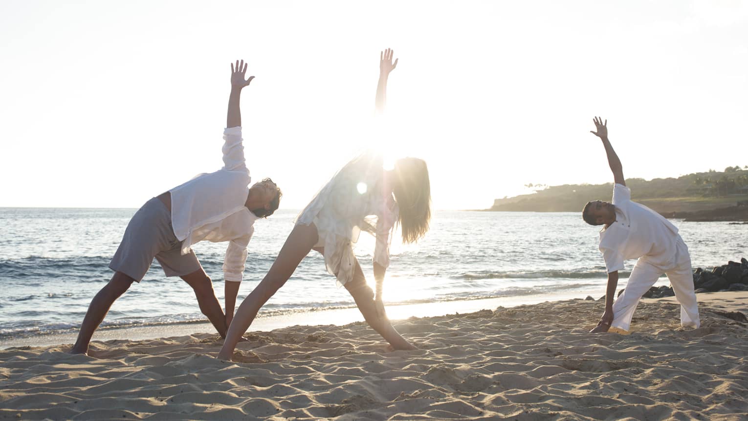 Three people stretch in yoga pose on sand beach at sunrise