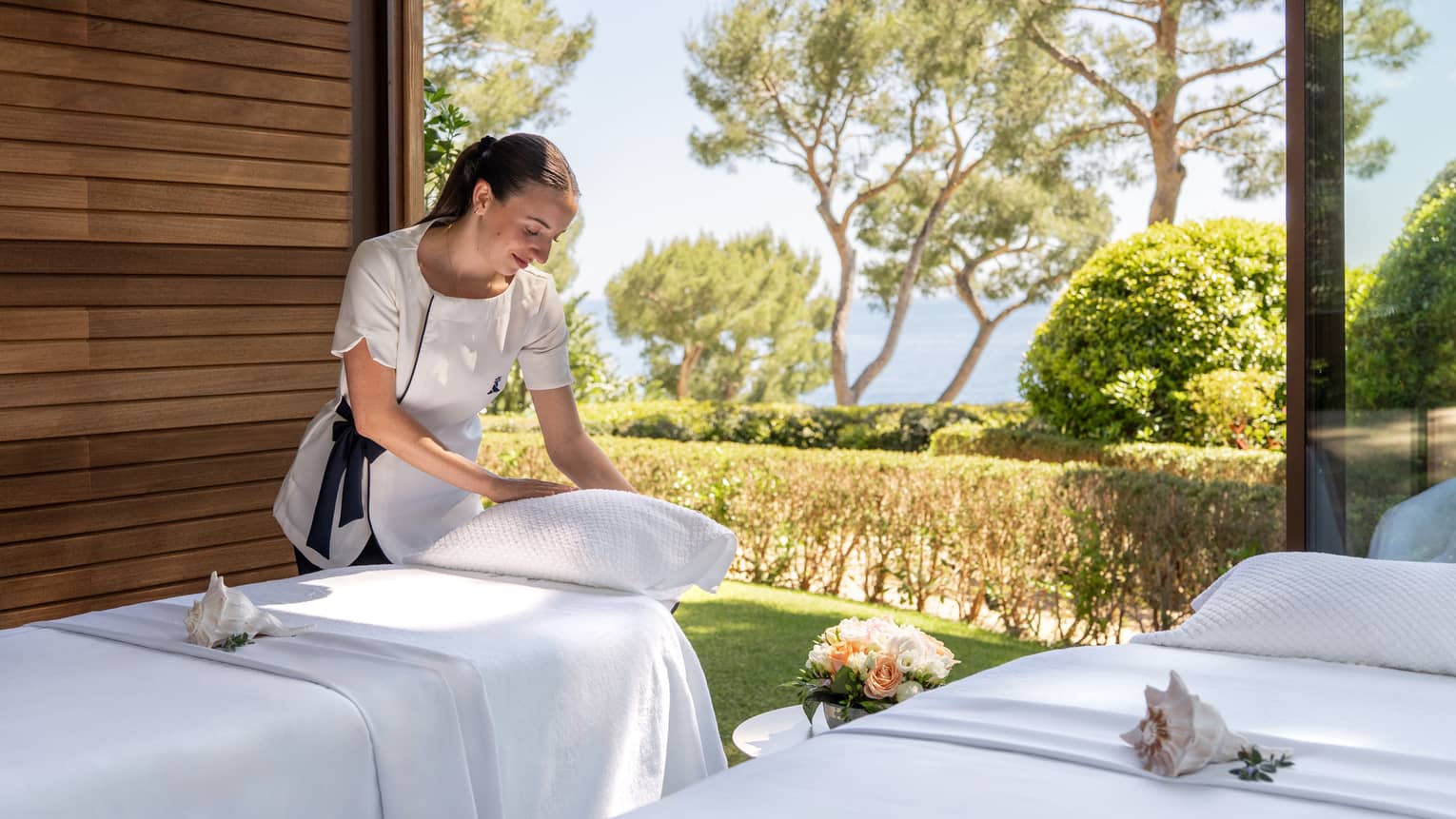 A naturally lit spa treatment space with two treatment tables in front of large glass windows. Trees and the sky are visible outside, and a person in a white uniform top places a pillow on one of the tables.