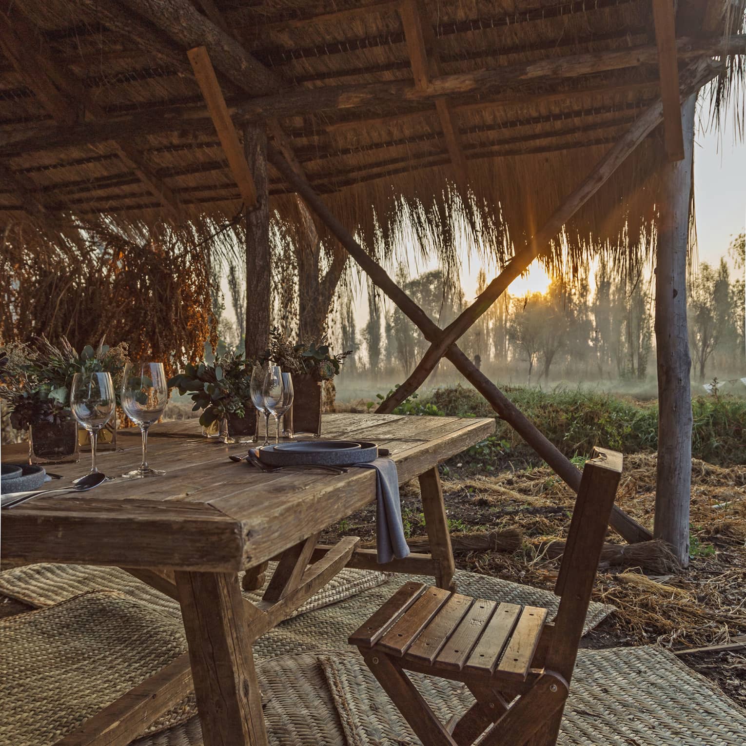The setting sun shining on a thatched wooden canopy covering a wooden table with rustic dinnerware and succulent plants.