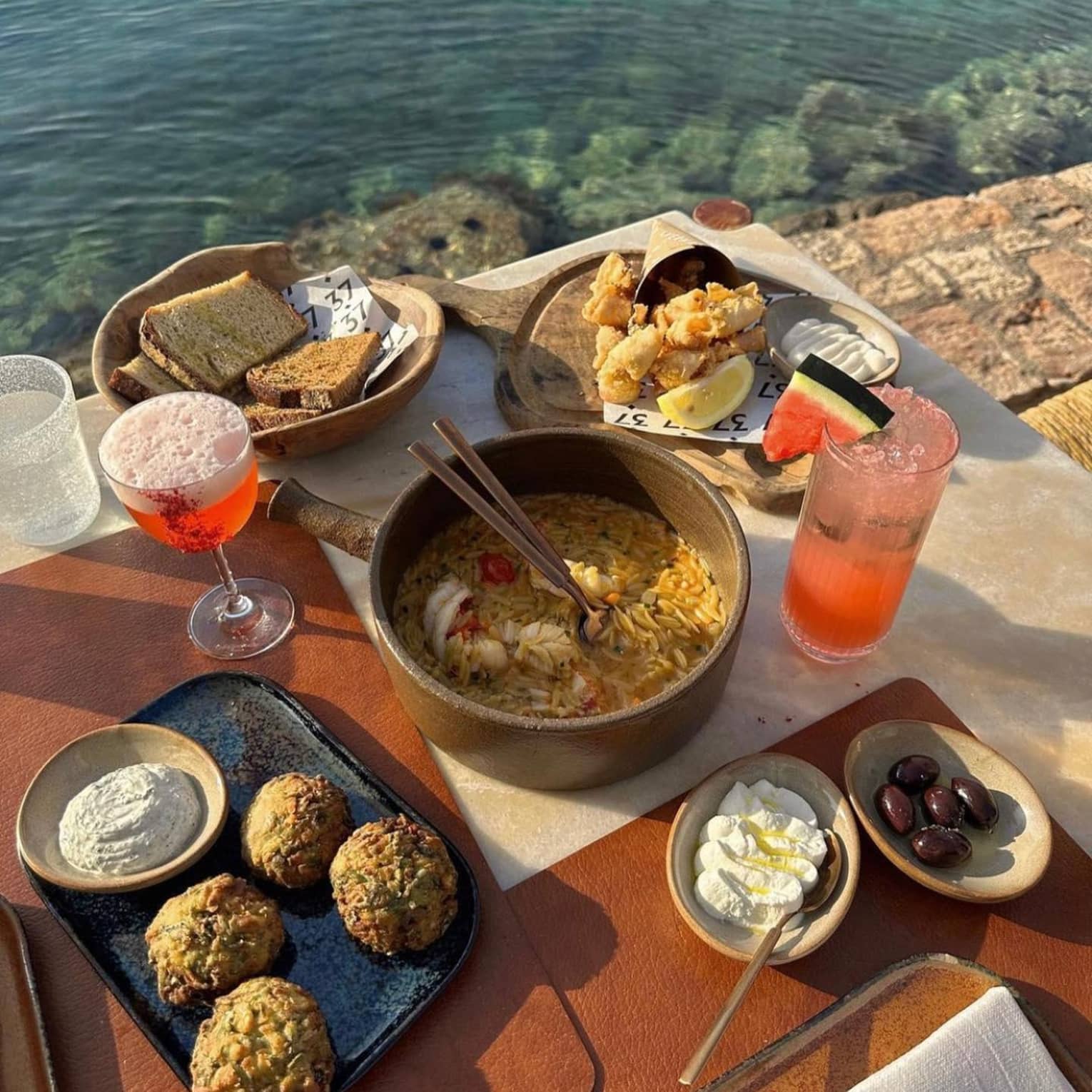 A wooden table right next to the sea, filled with various fresh food dishes, and colourful cocktails