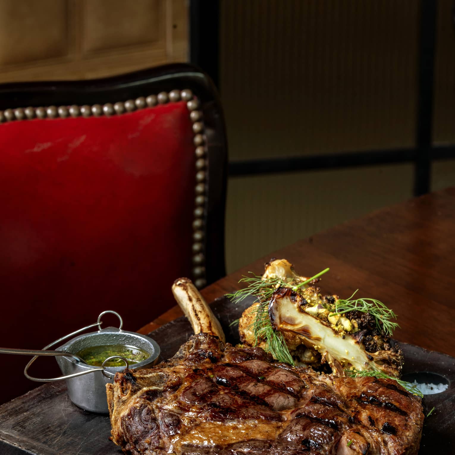 A thick, succulent cowboy ribeye steak rests on a wooden serving board, flanked by grilled vegetables and chimichurri sauce.