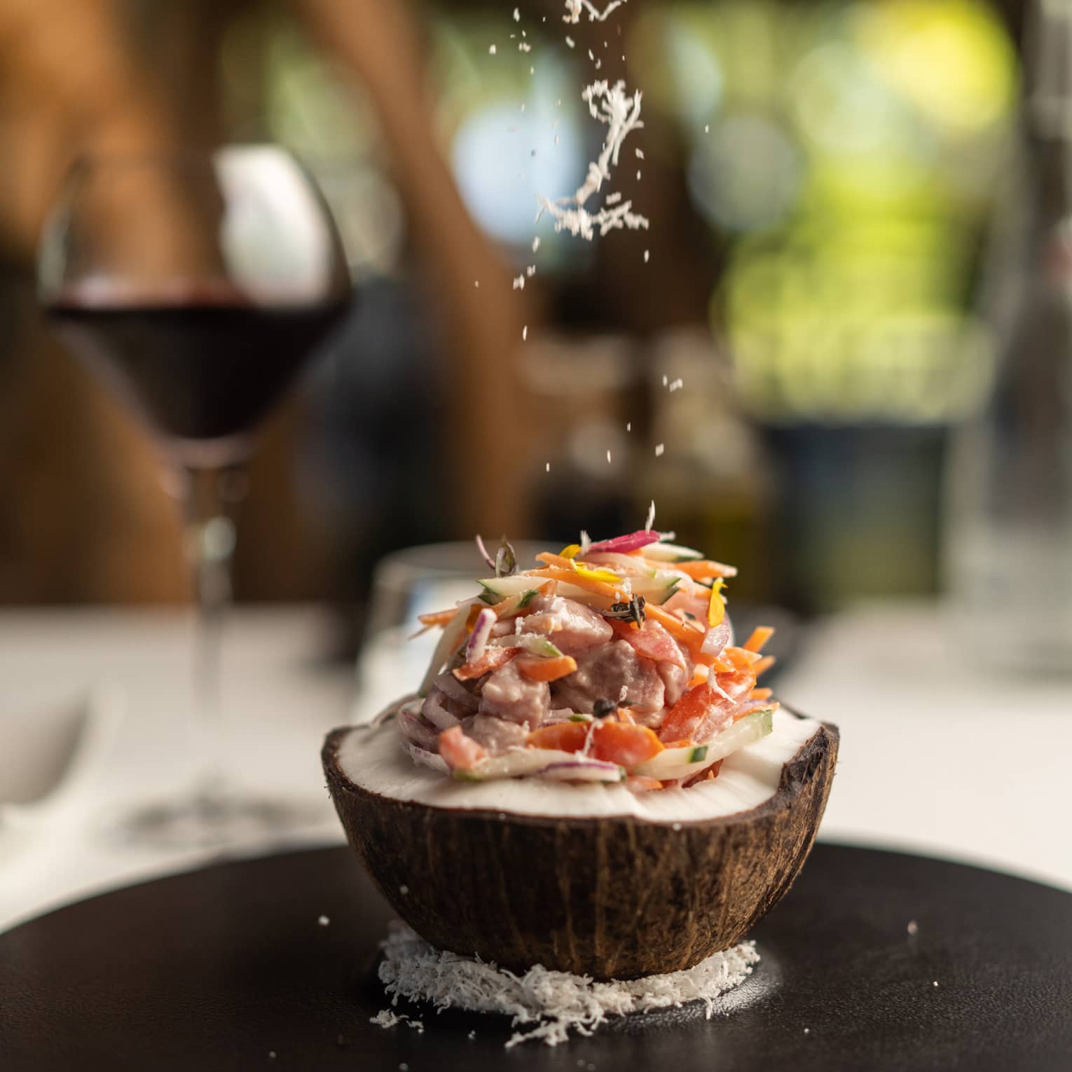 Close-up of an open coconut filled with fish and thinly sliced vegetables, atop a bed of coconut flakes on a round platter.
