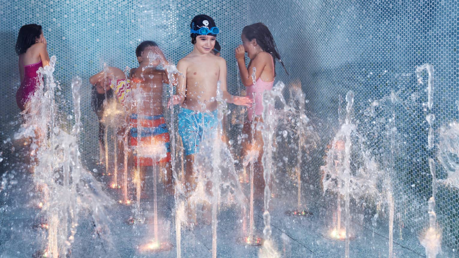 Six children play in a blue-tiled splash pad as water rises up from different holes in the floor