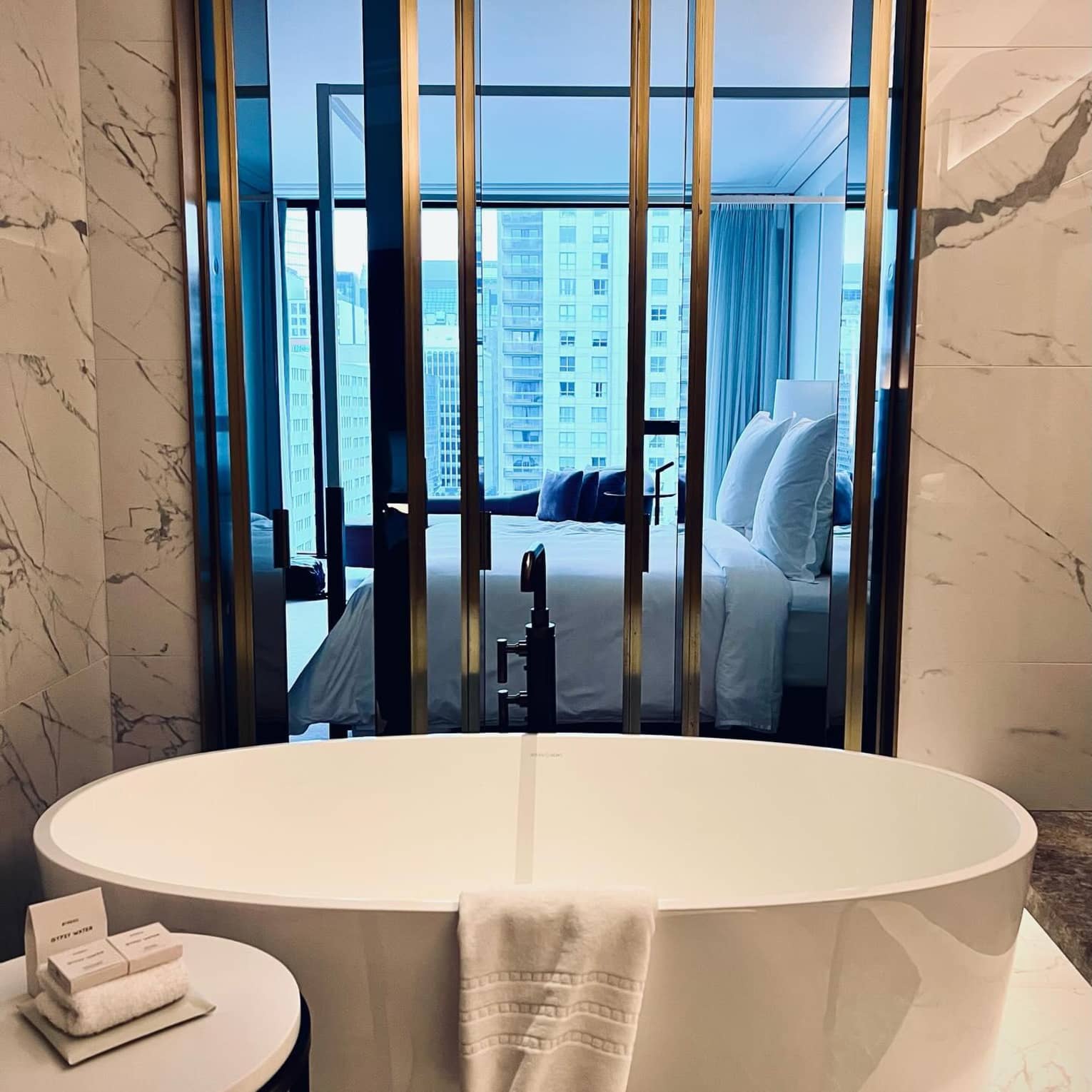 A spacious garden bathtub surrounded by marbled floors and walls overlooking a modern Four Seasons suite with a view of the Montreal skyline