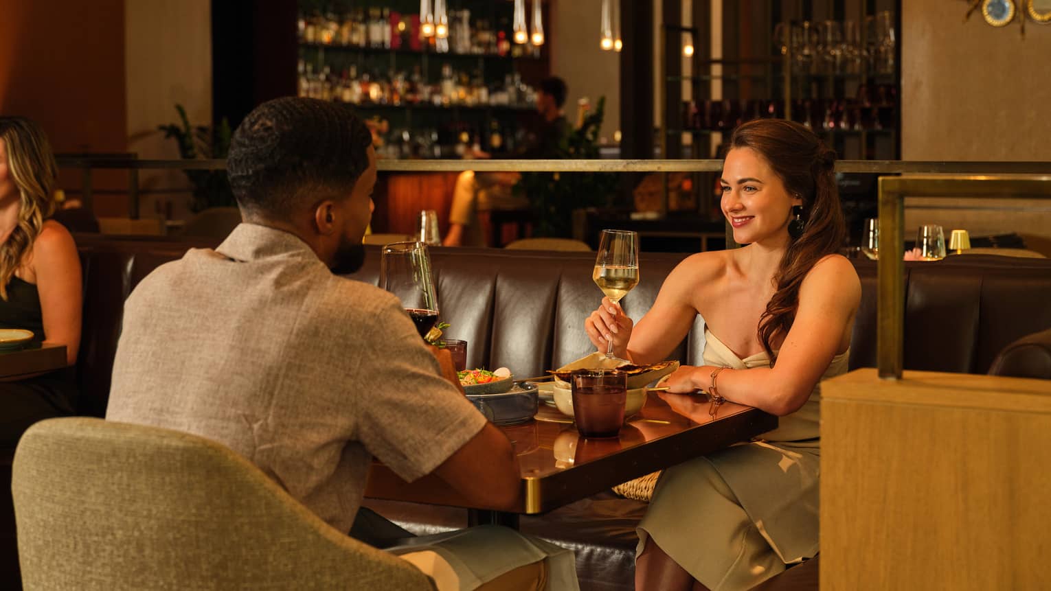 Two people drink beverages from stemmed glasses, sitting across from each other at a dining table in a serene and cozy indoor dining space.