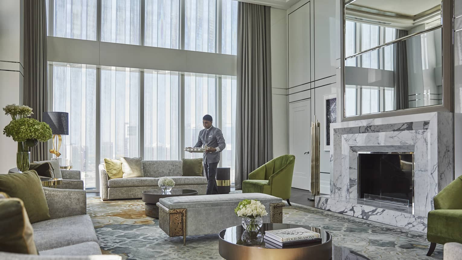 Luxurious hotel living room with high ceilings, marble fireplace, plush gray sofas, green accent chairs, floral arrangements and an attendant serving refreshments