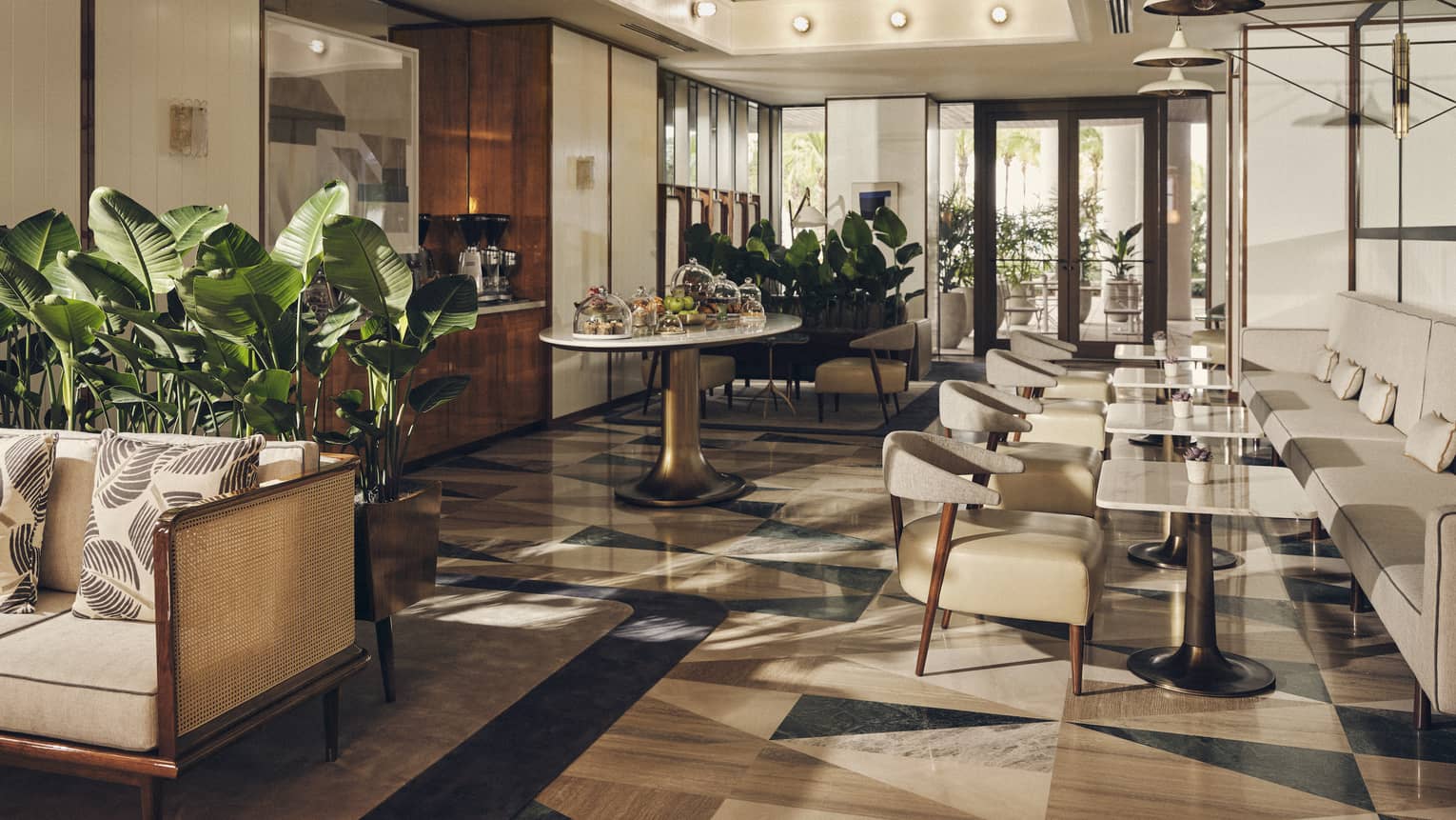 A dining area with a long white booth and white chairs, tile floor, plants and sofa.