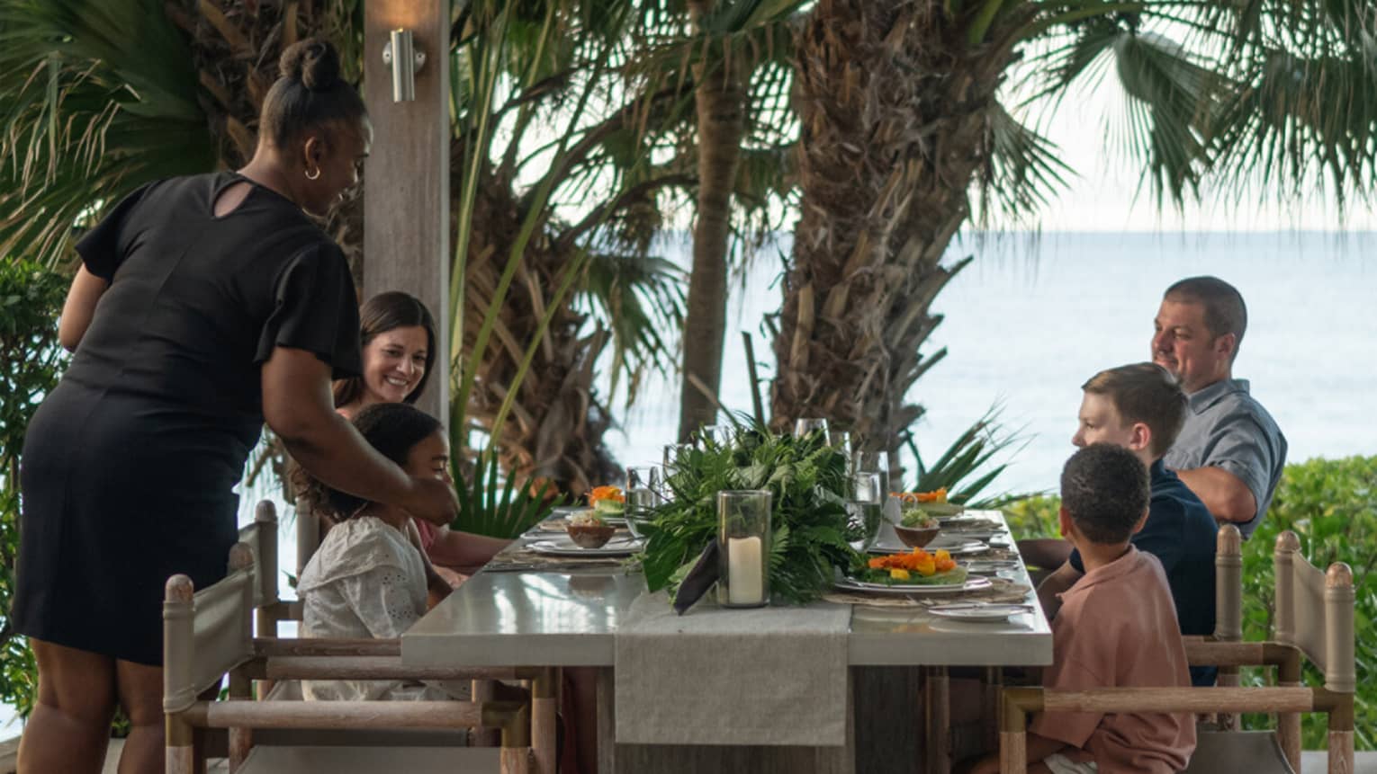 Group of people enjoying an outdoor meal at a tropical seaside dining table.