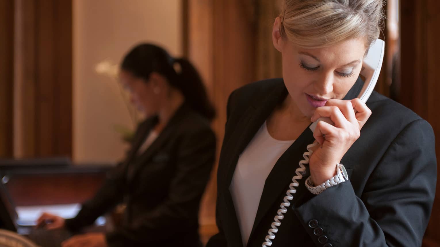 Close-up of woman in dark suit jacket holding telephone, cord, silhouette of hotel reception staff in background