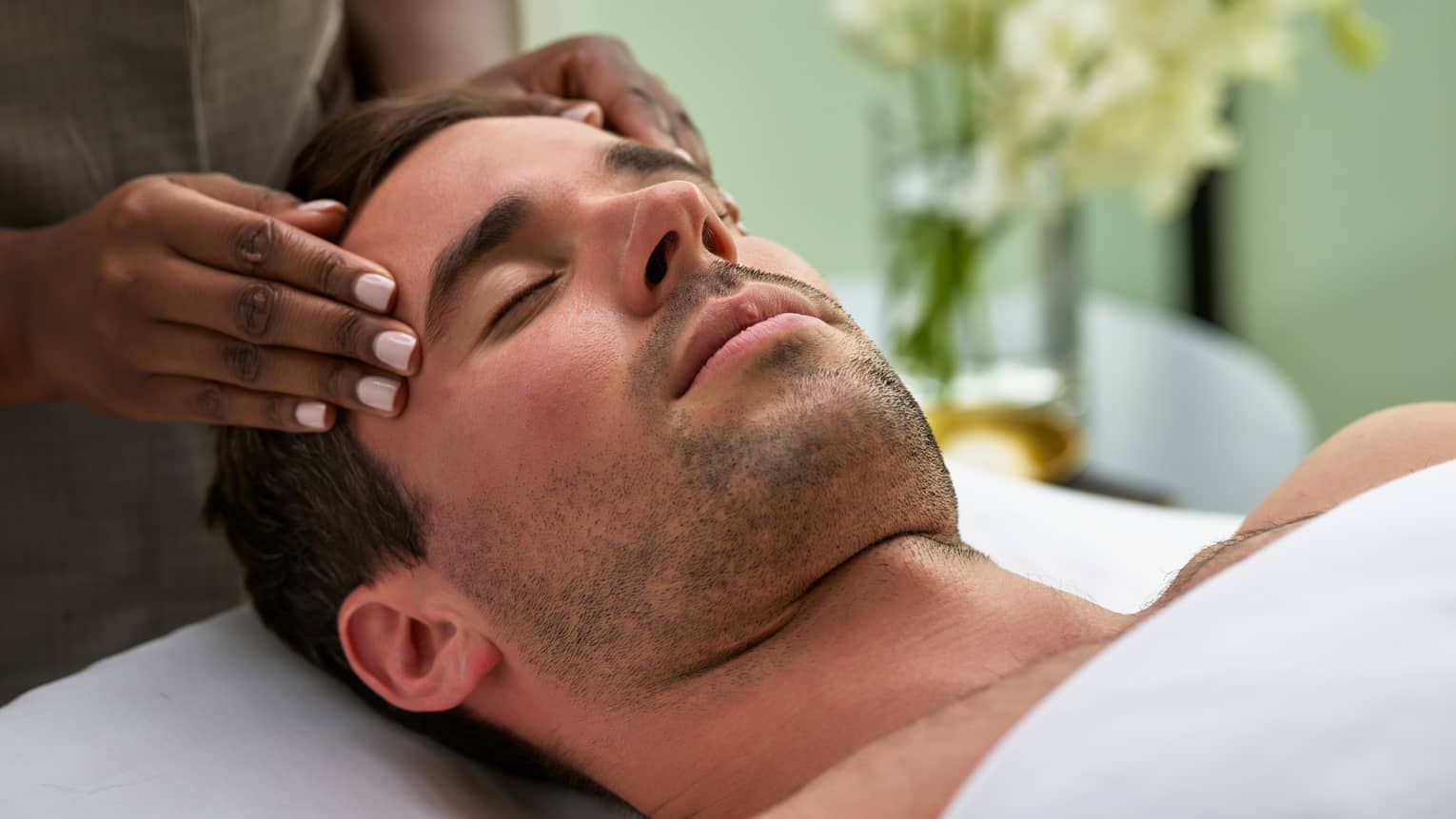 Man lies on massage table with eyes closed as hands massage his temples, forehead
