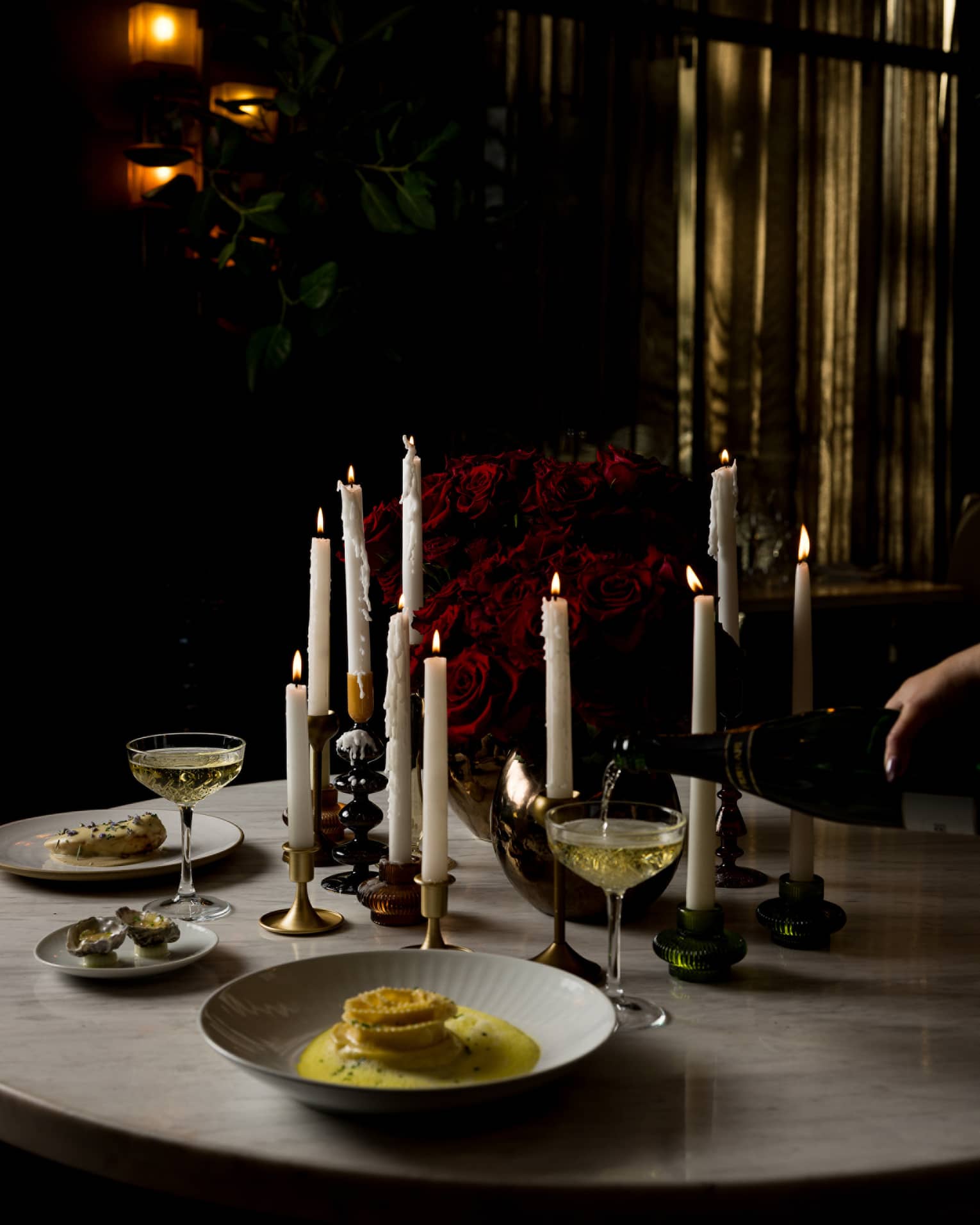 A round marble table topped with various pillar candles, a red rose centrepiece and food and drink for two is set in a dimly lit room
