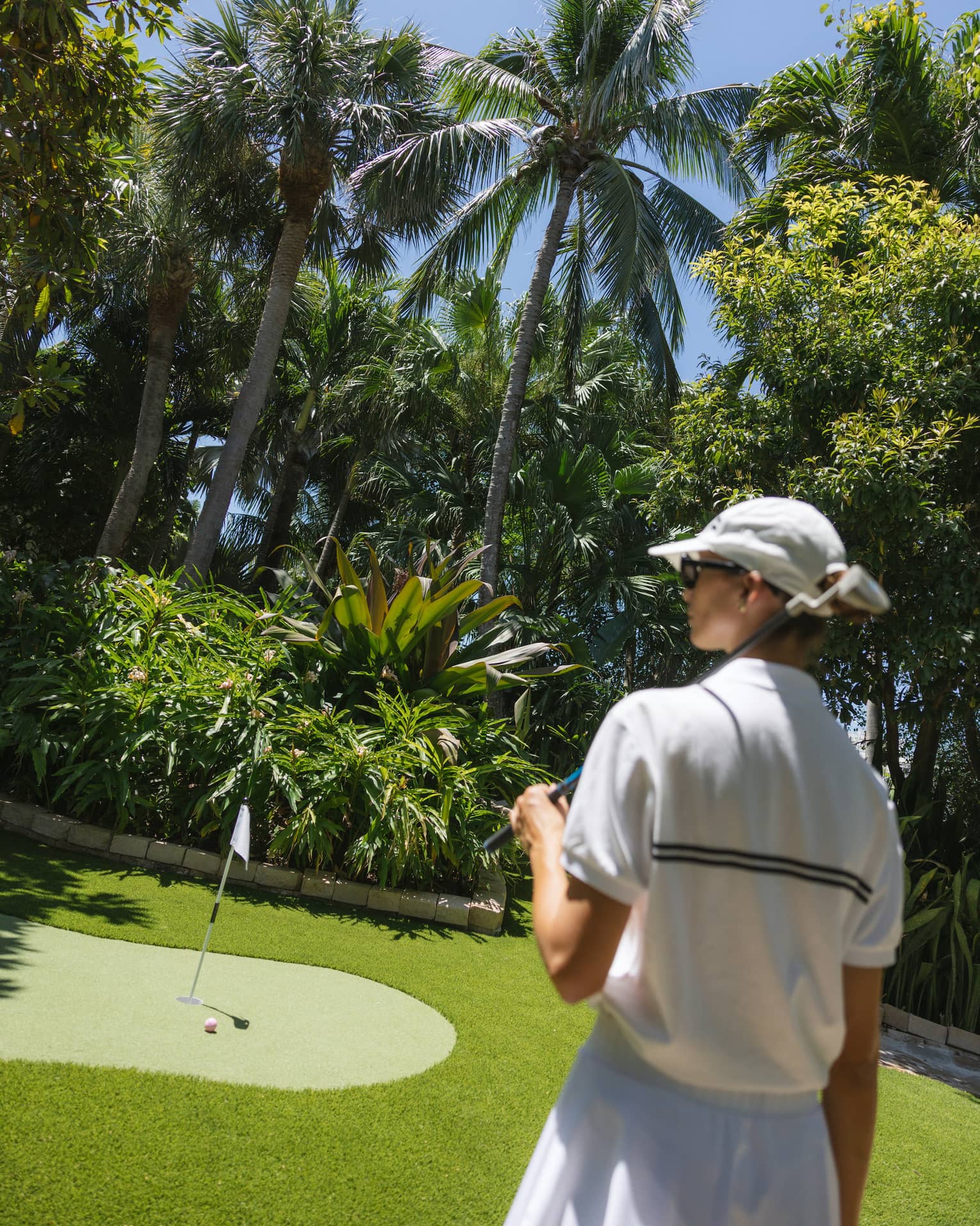 Golfer wearing white sweater vest, white skirt and white hat walks toward a putting green surrounded by palm trees
