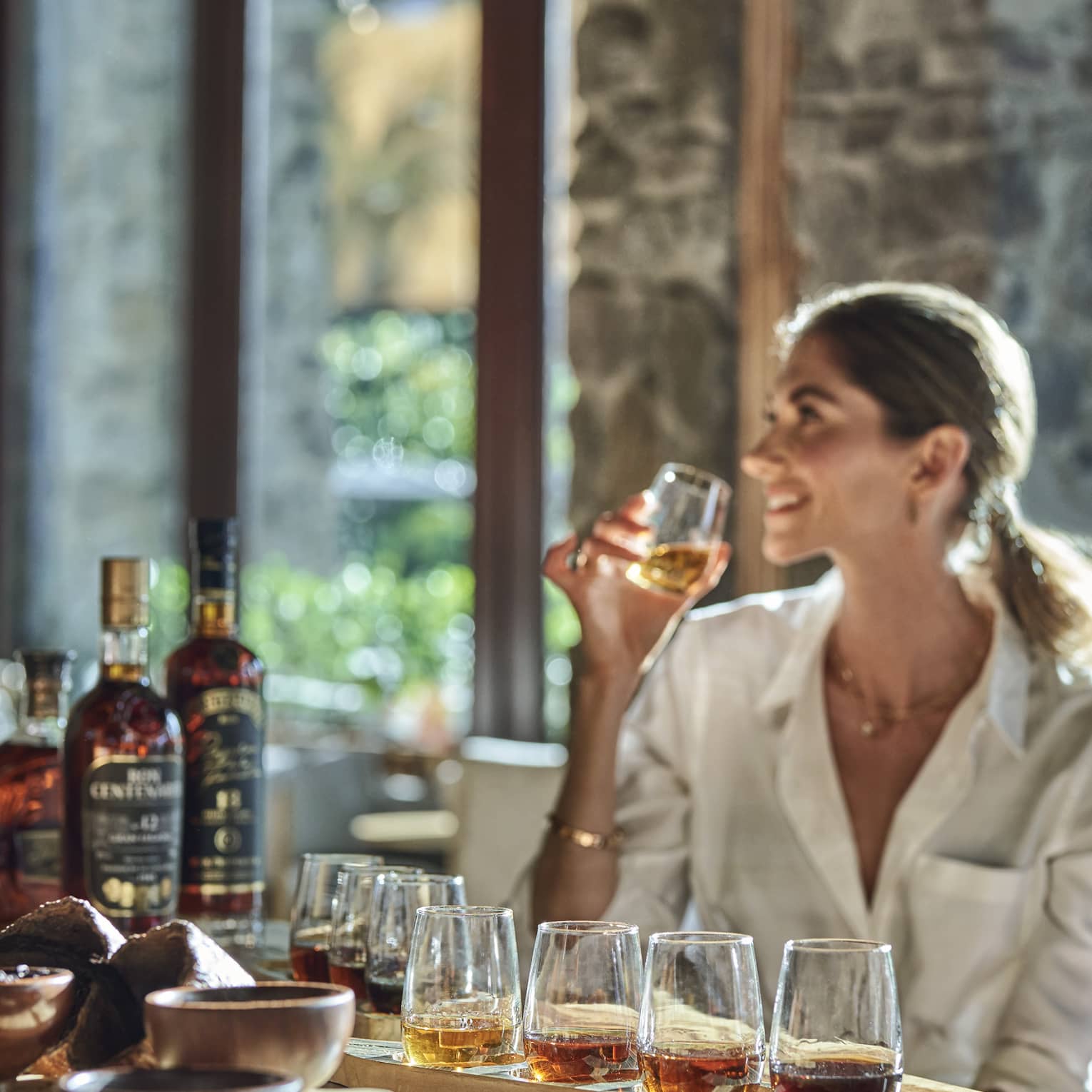 A guest smiles while holding a glass from a flight of eight rums, sun streaming in from behind through a bank of windows.