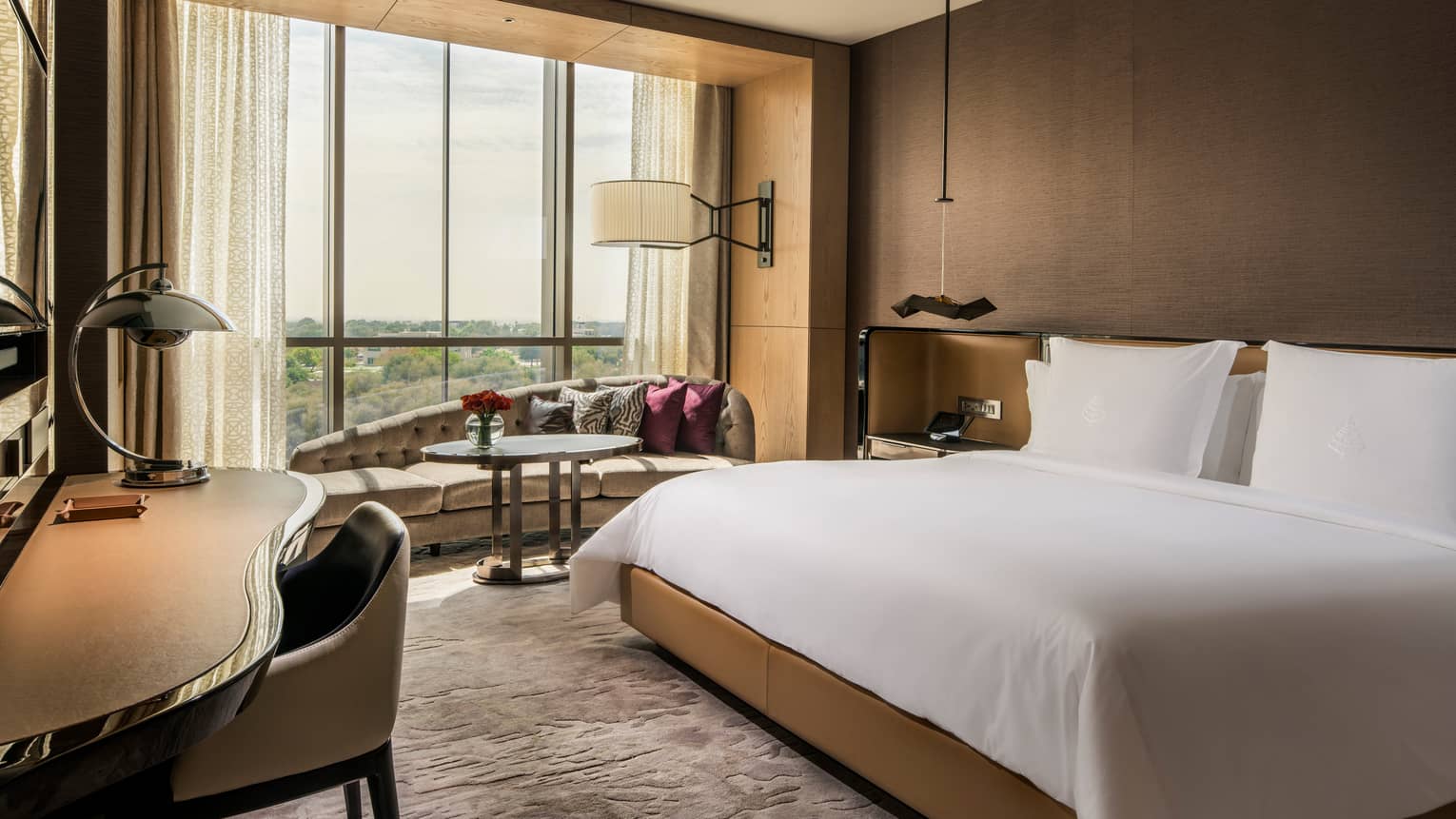 King-sized hotel bed with white linens, modern sofa under sunny window, desk and chair
