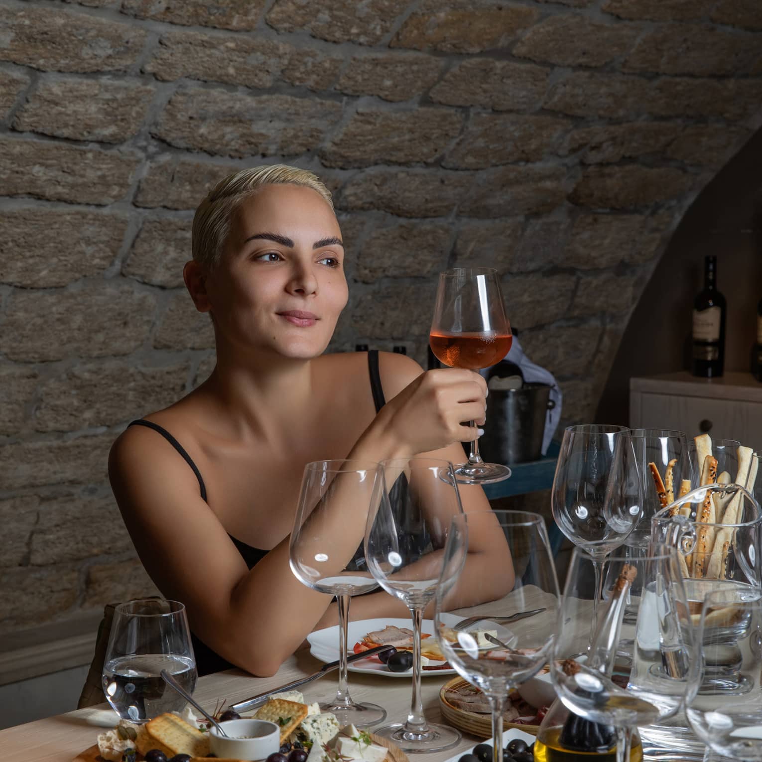 At a table set with glassware and hors-d’oeuvres, a wine enthusiast holds a glass containing a rose-coloured beverage.