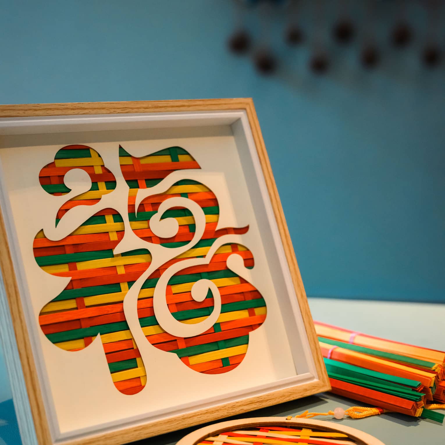 Coloured strips of straw woven together in wooden frames, creating an art pattern by itself or placed behind a stencil.