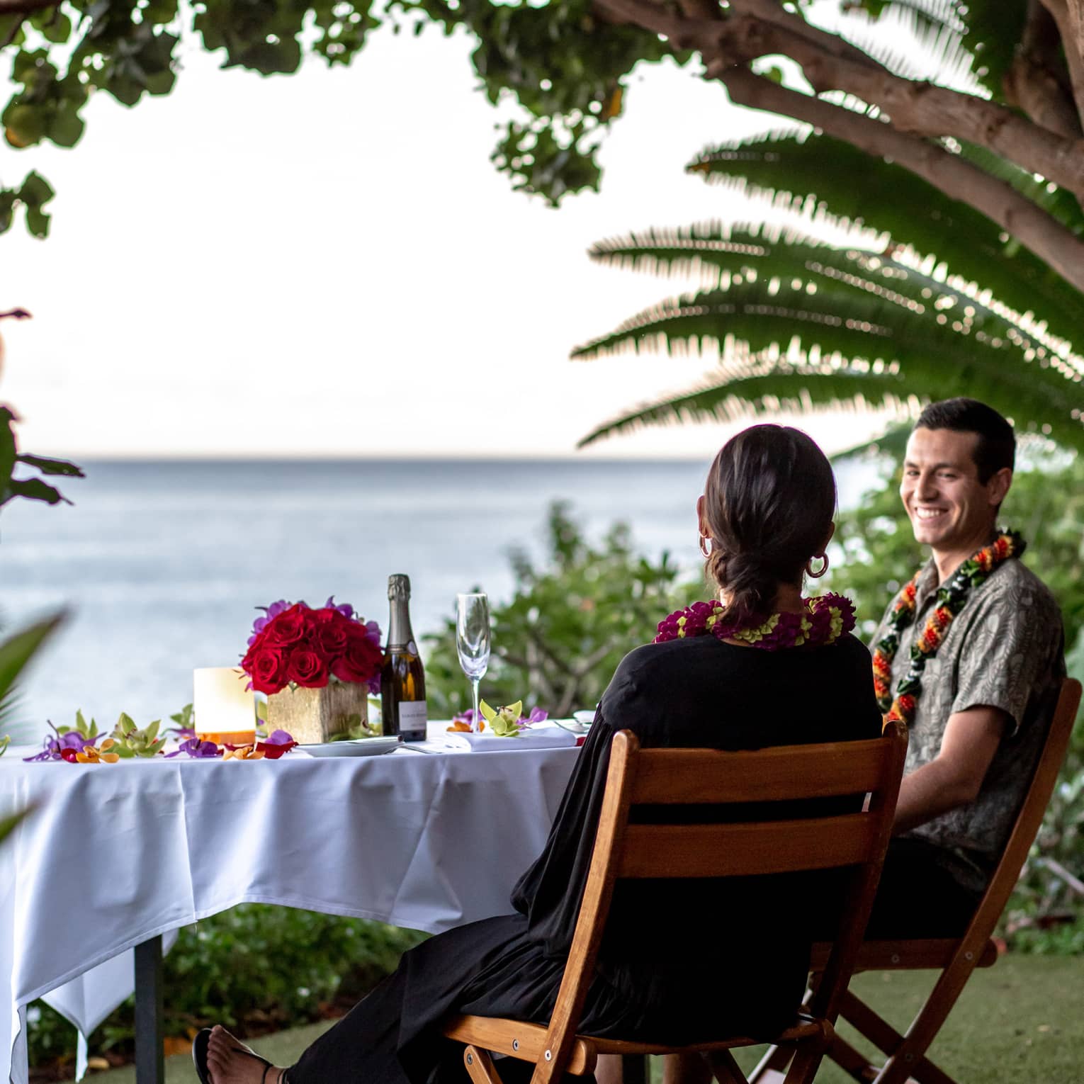 Under lush trees, a smiling couple sits at an ocean-view table set with a centrepiece of red roses and a bottle of champagne.