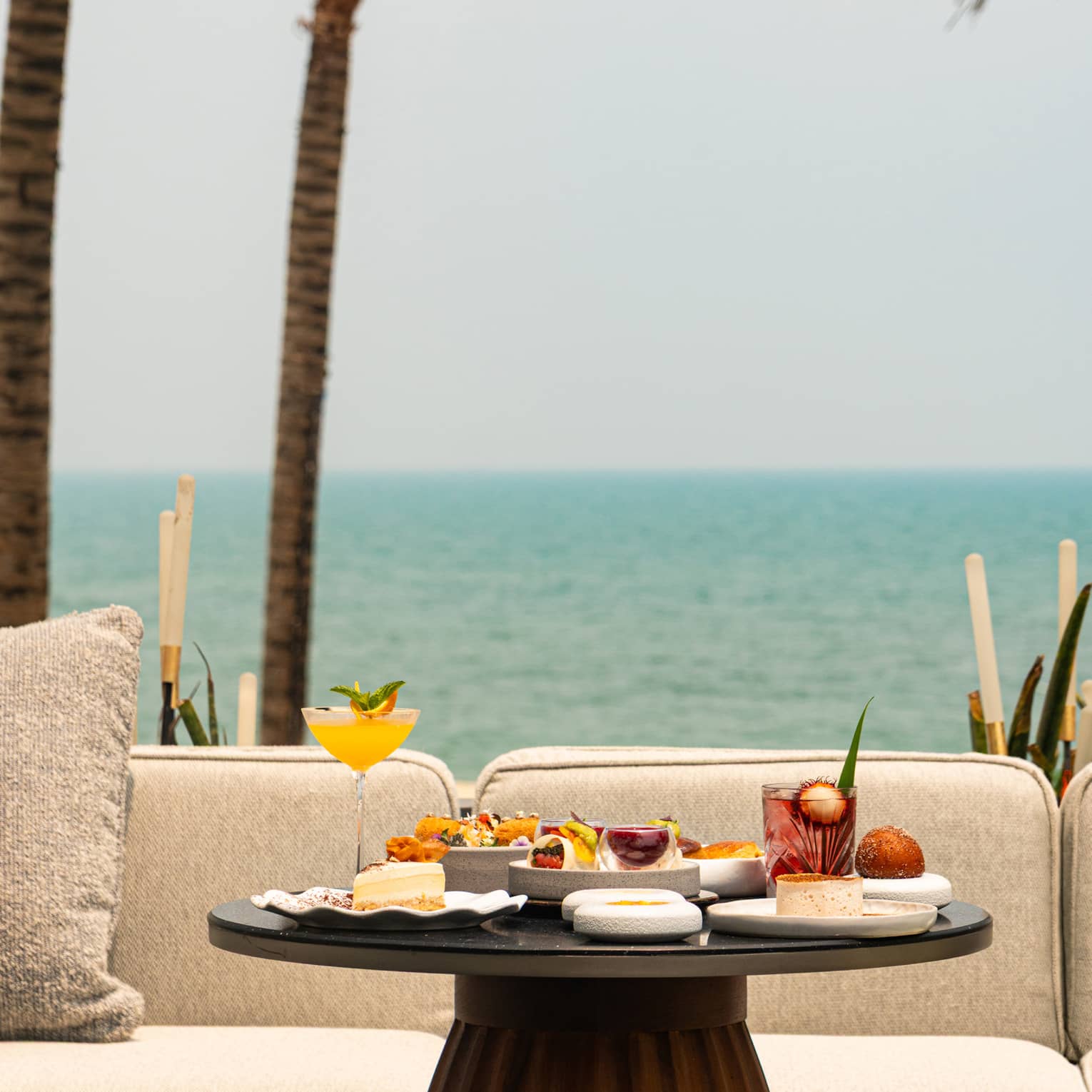 Round table set for afternoon tea in front of cream outdoor sofa, overlooking the sea