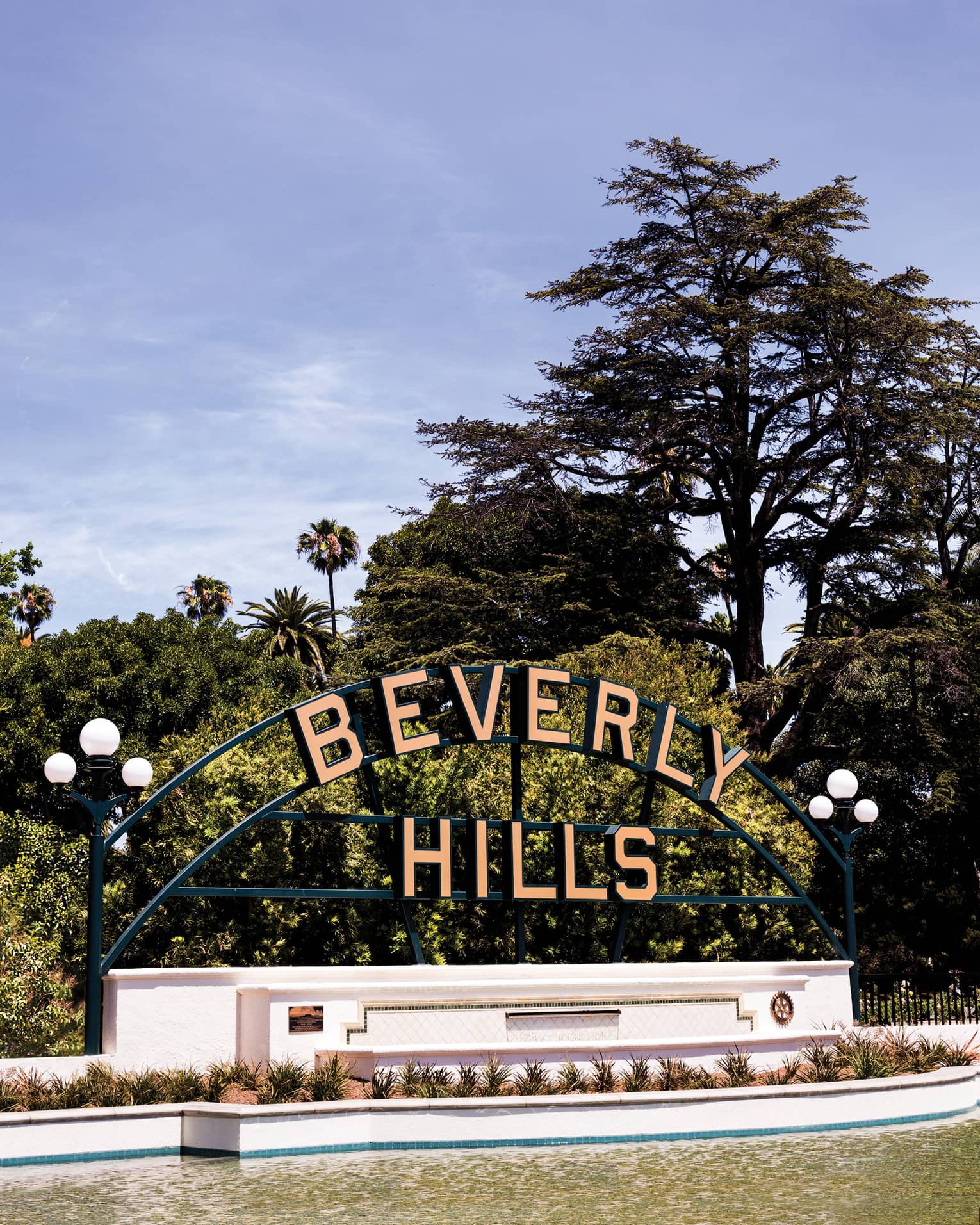 Large pink letters, mounted on a metal frame, spell out the name "Beverly Hills" against an outdoor backdrop of leafy trees.