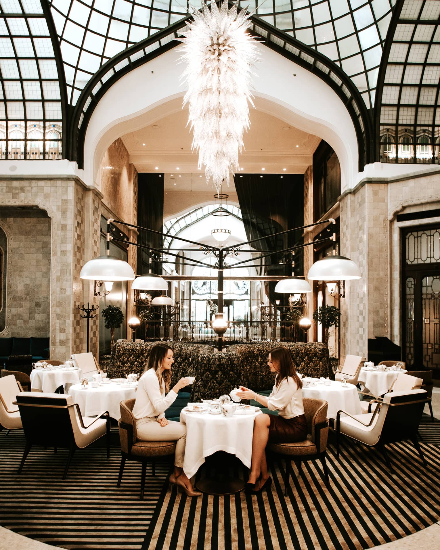Elegant dining area with a grand chandelier, high arched glass ceiling and modern seating, where two people are enjoying tea at a small table
