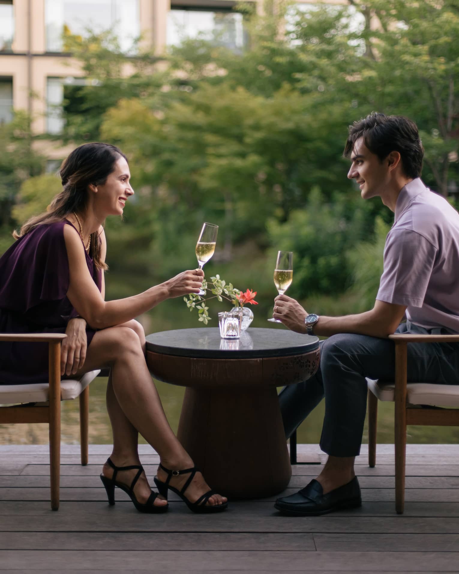 Two people sitting outdoors, toasting with champagne glasses at a small table with a flower vase