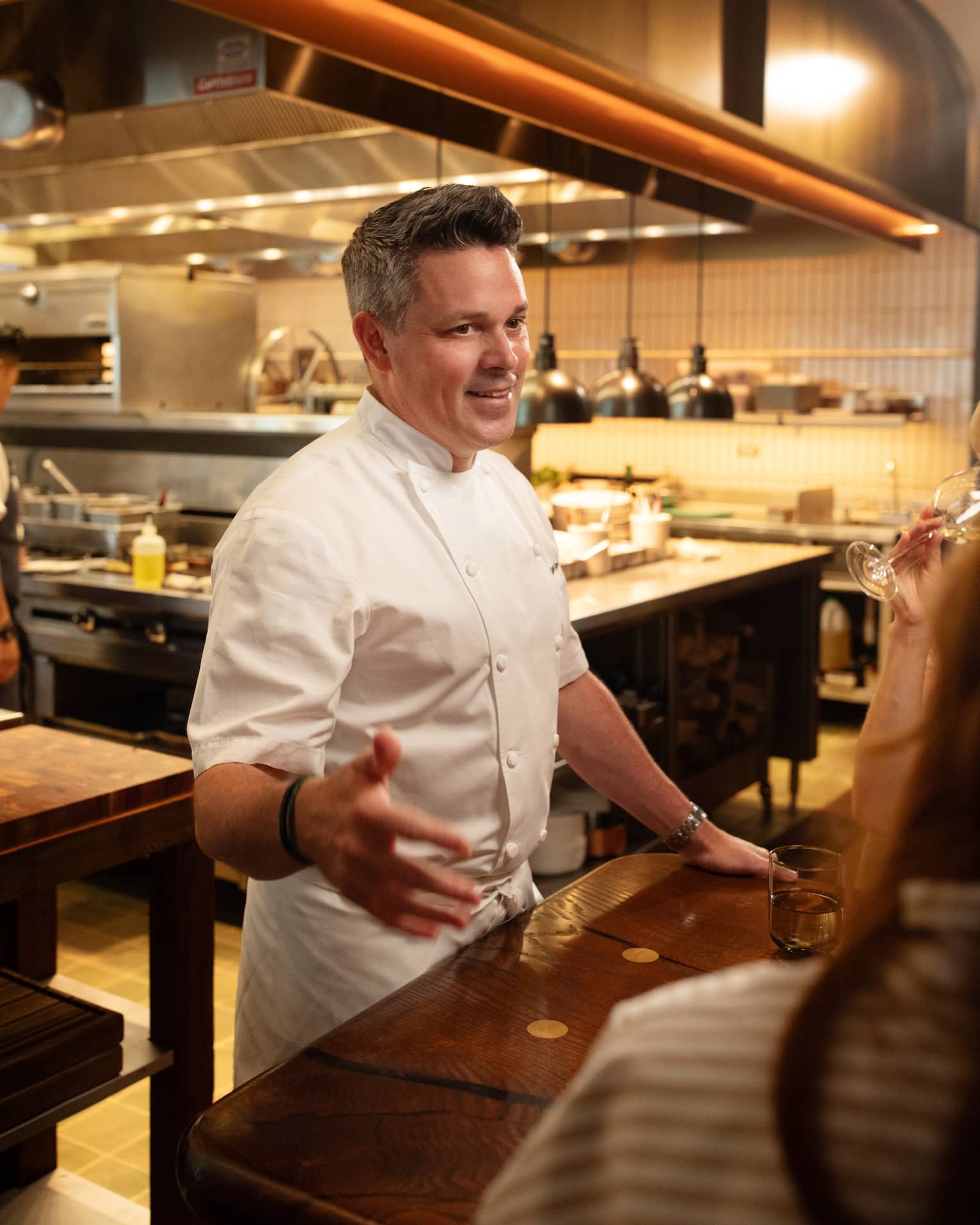 Chef standing beside a dining table in the kitchen talking to guests.
