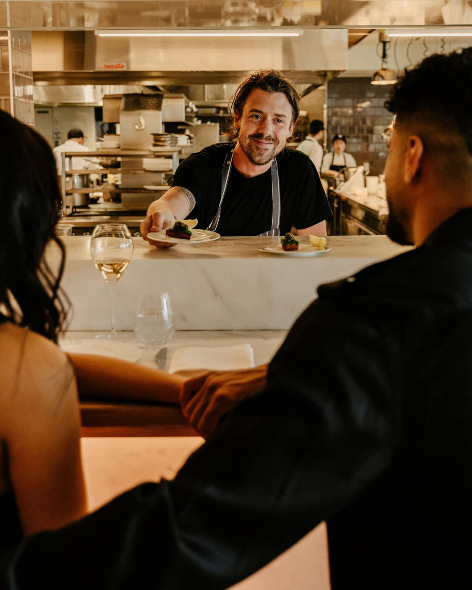 Rear view of a couple sitting at a counter as a smiling chef hands them a small plate, a blurred kitchen in the background.