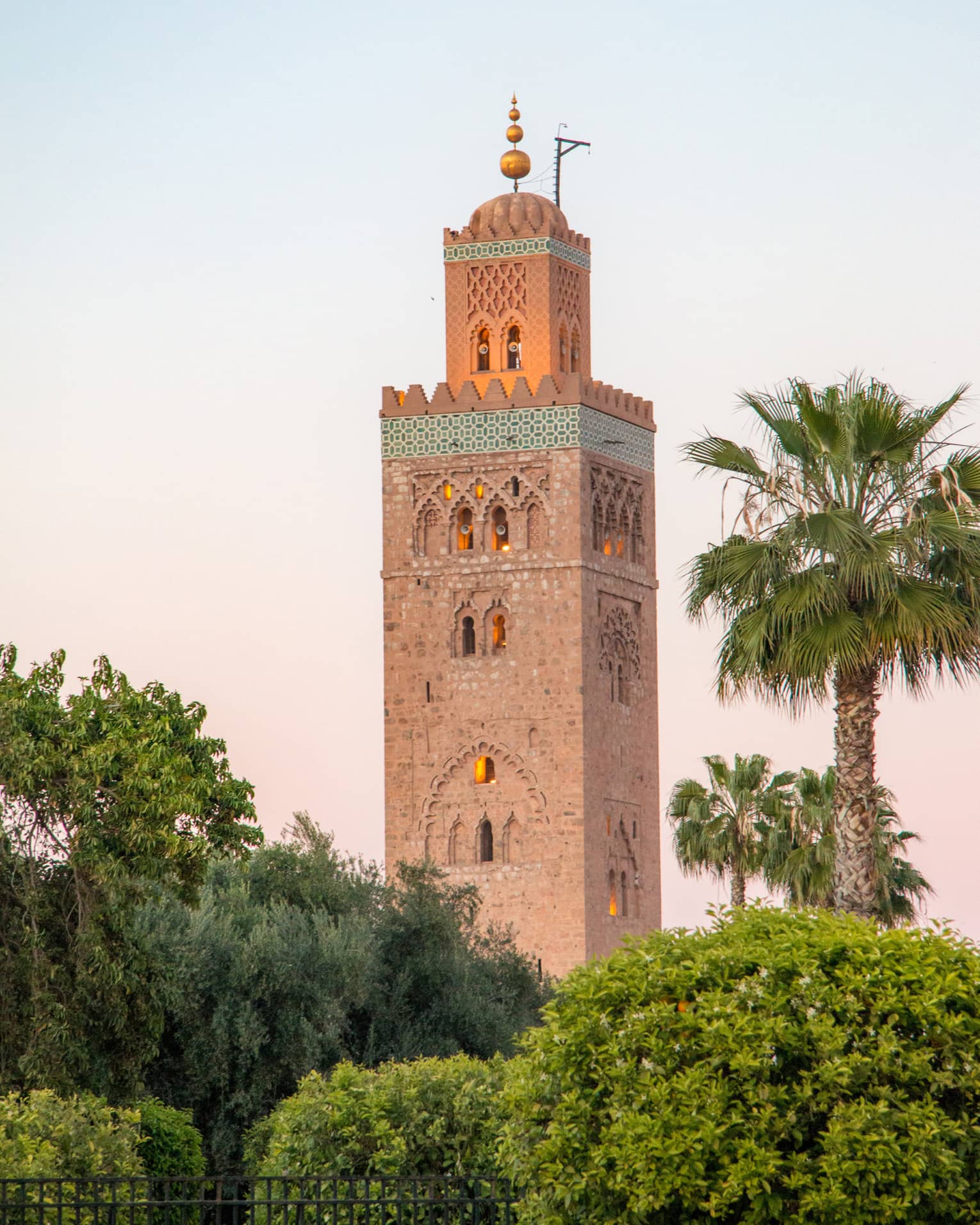Rising from amid bushes and palms, a towering minaret with small domed archway windows, golden light emanating from within.