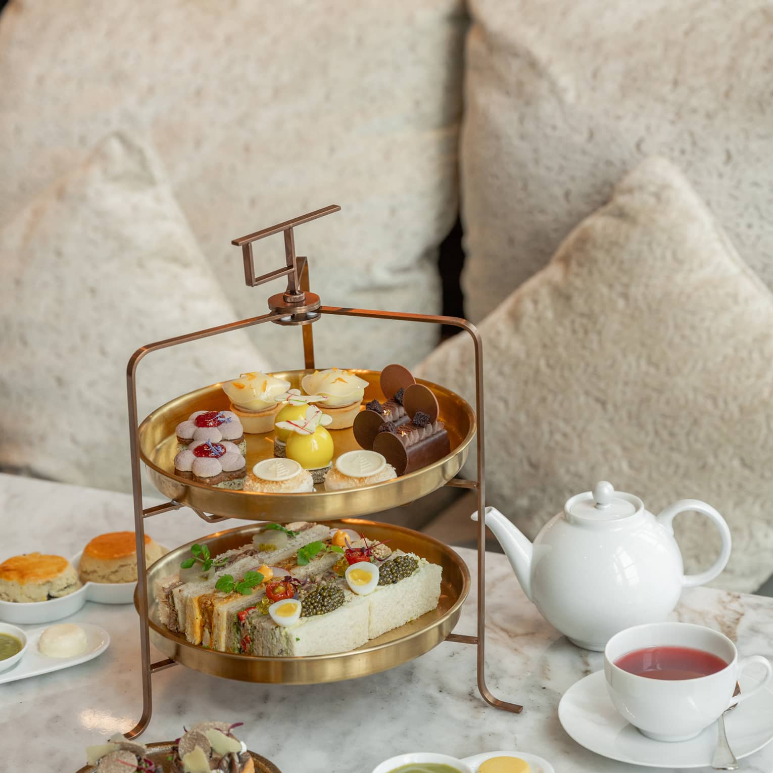 Two-tiered afternoon tea spread on marble coffee table in front of tan sofa