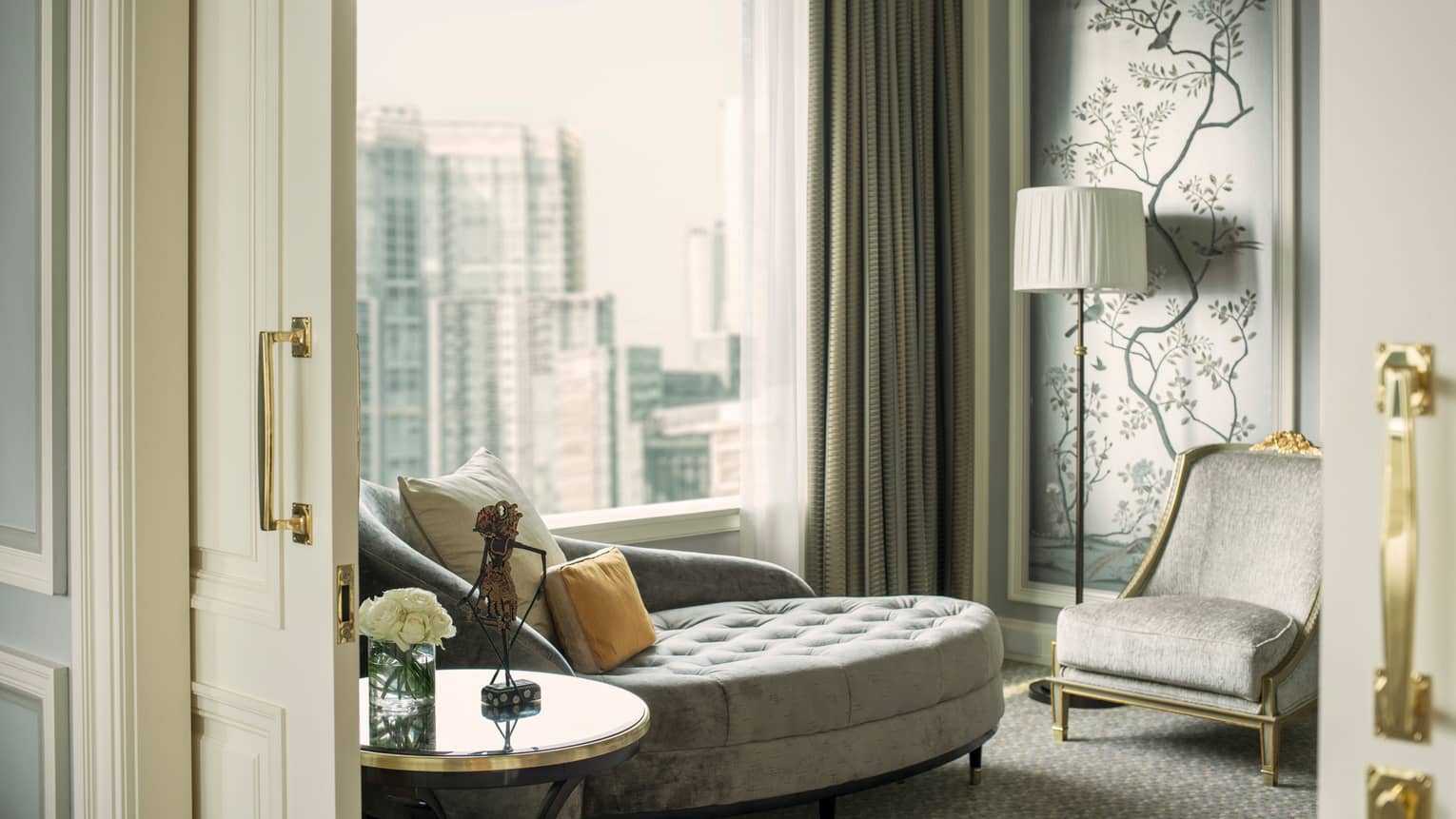 Round grey velvet chaise in front of sunny floor-to-ceiling window, near wall panel with trees, flowers and armchair