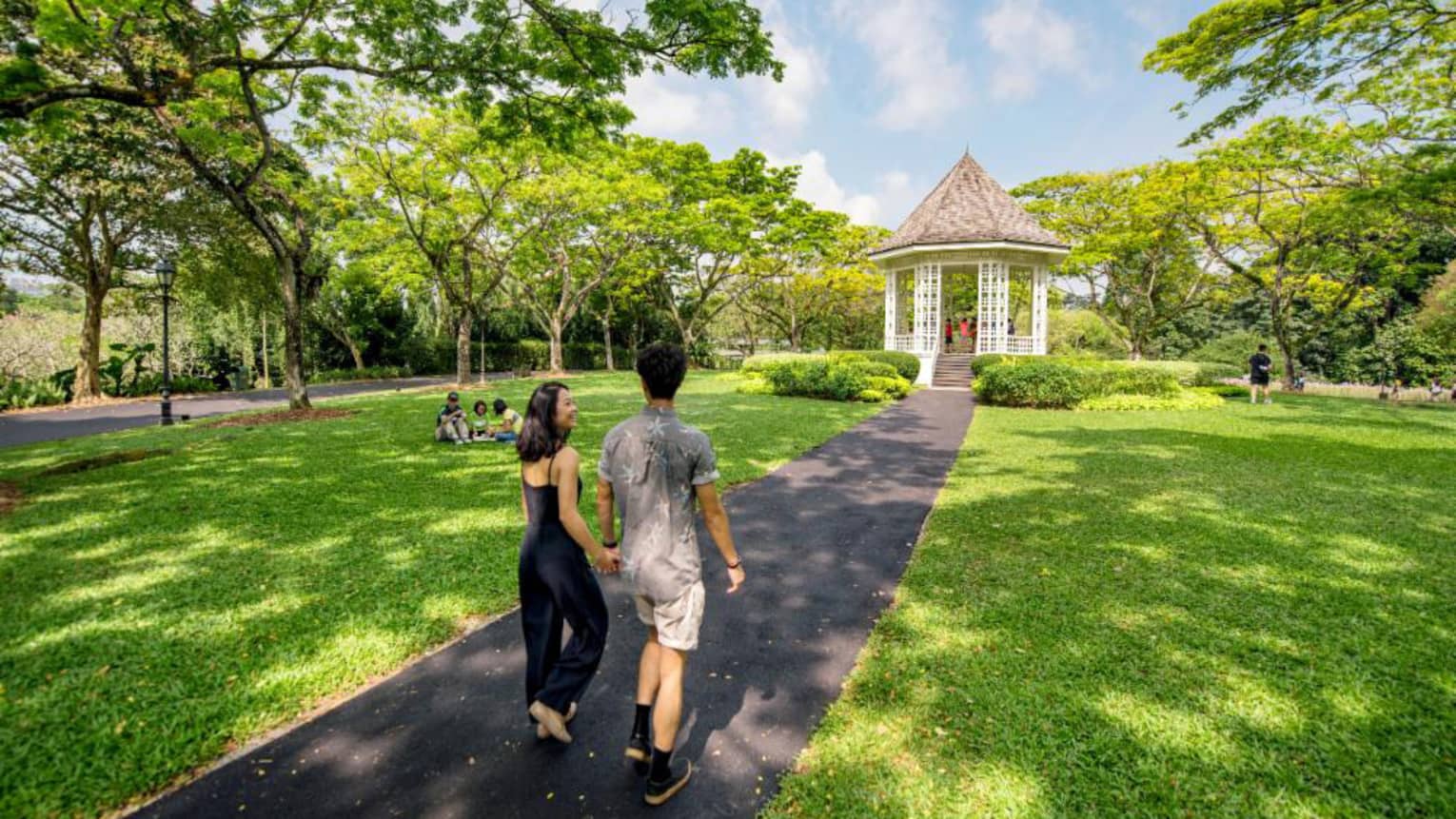 A man and woman hold hands and walk along an asphalt trail through the Singapore Botanic Gardens