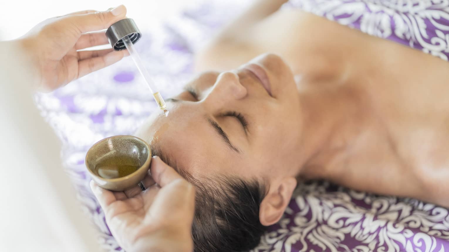 A woman receiving a relaxing facial treatment, with oil being applied from a dropper, lying on a patterned purple sheet.