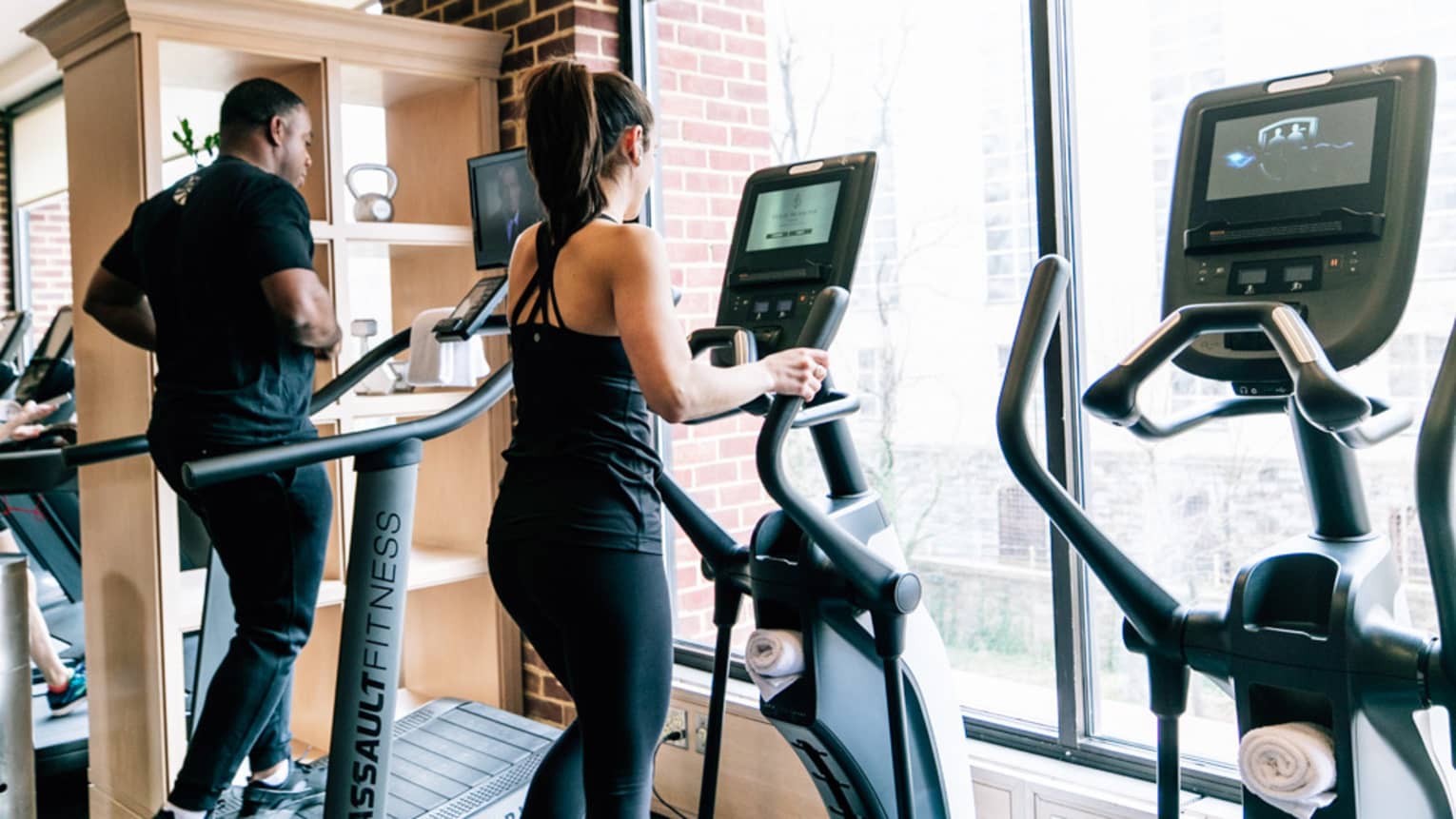 Woman and man on elliptical trainers at the gym, facing large window