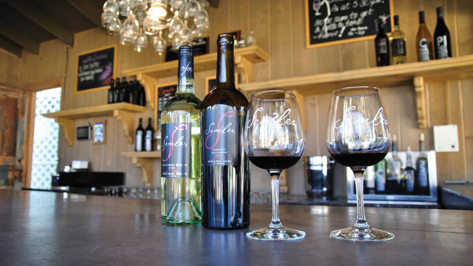 A red and white bottle of wine next to two glasses of red wine. More bottles are on shelves in the back.