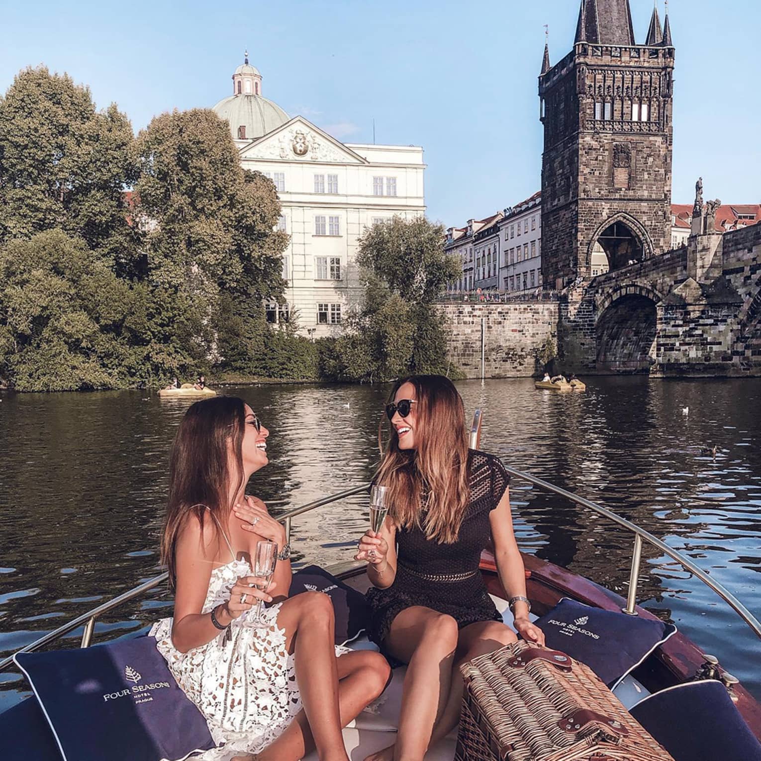 Two people sitting on a boat, enjoying champagne, with a wicker picnic basket and historic buildings and a bridge in the background.