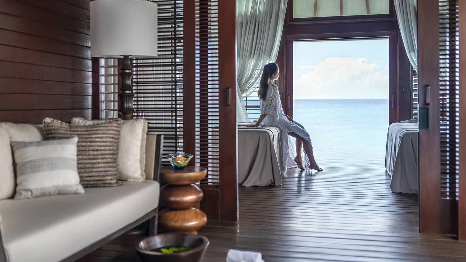 Person sitting on a massage bed in a serene spa treatment room with ocean view, surrounded by cozy seating and wooden décor
