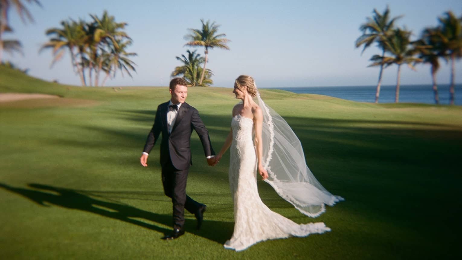 A man in a black tuxedo and a woman in a white wedding gown and veil walk hand in hand along a lush green lawn surrounded by palm trees and backed by clear blue sky and an ocean horizon.