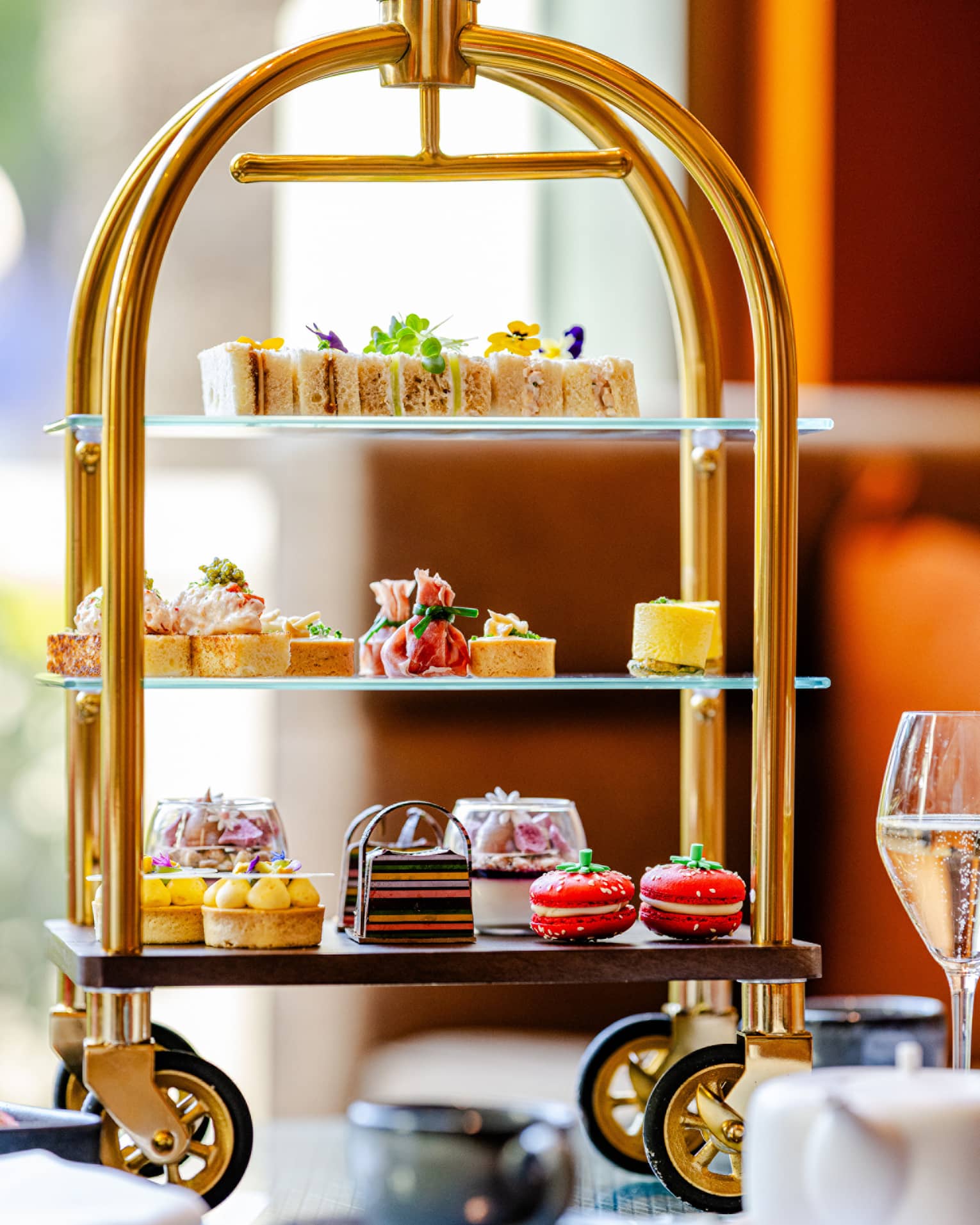 A tiered serving tray on small wheels holds an assortment of colourful small bites. It sits on a tabletop next to a stemmed glass, a teapot, and a teacup and saucer.
