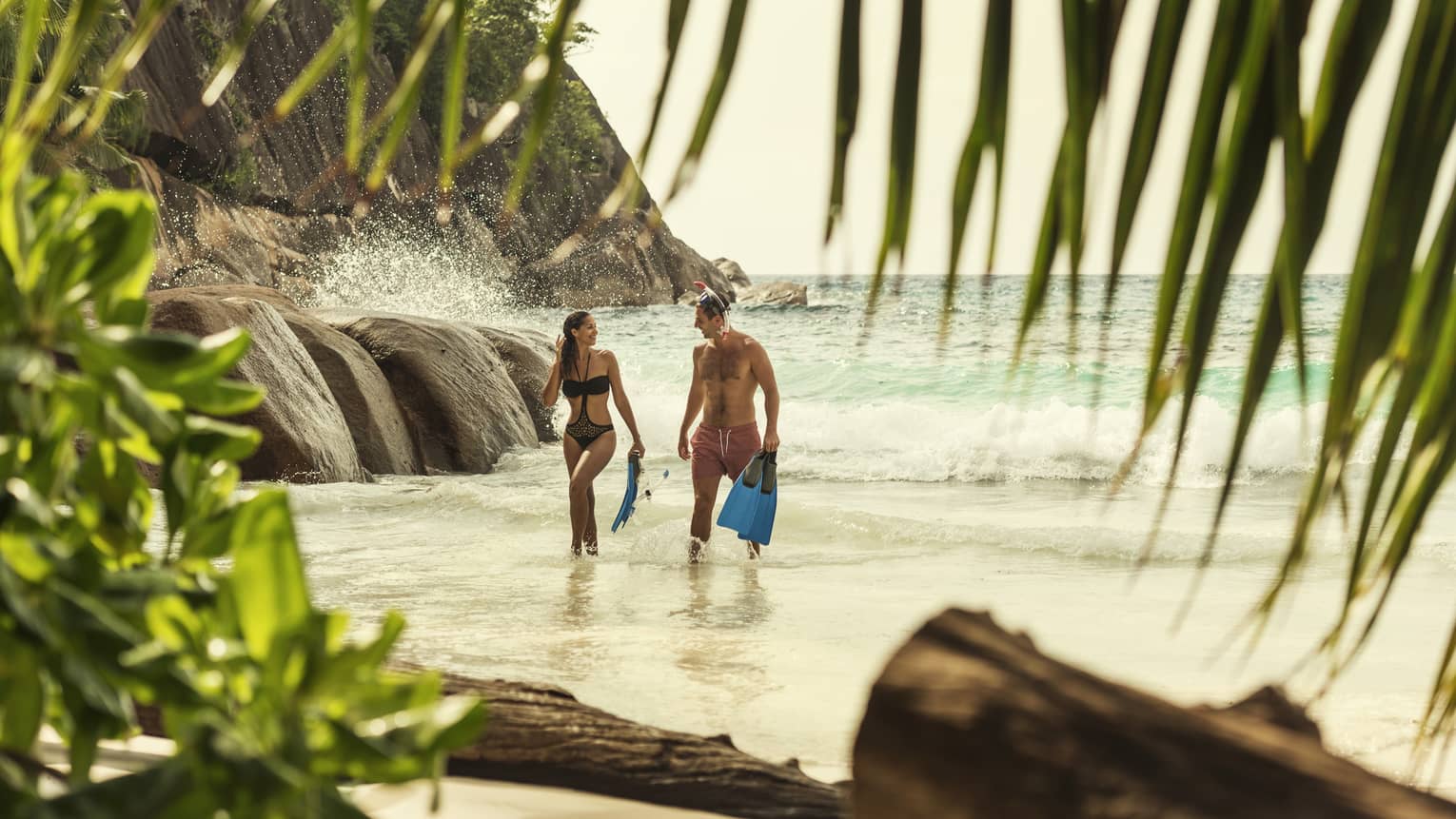 Man and woman in swimsuits hold snorkelling fins, walk from the ocean to beach