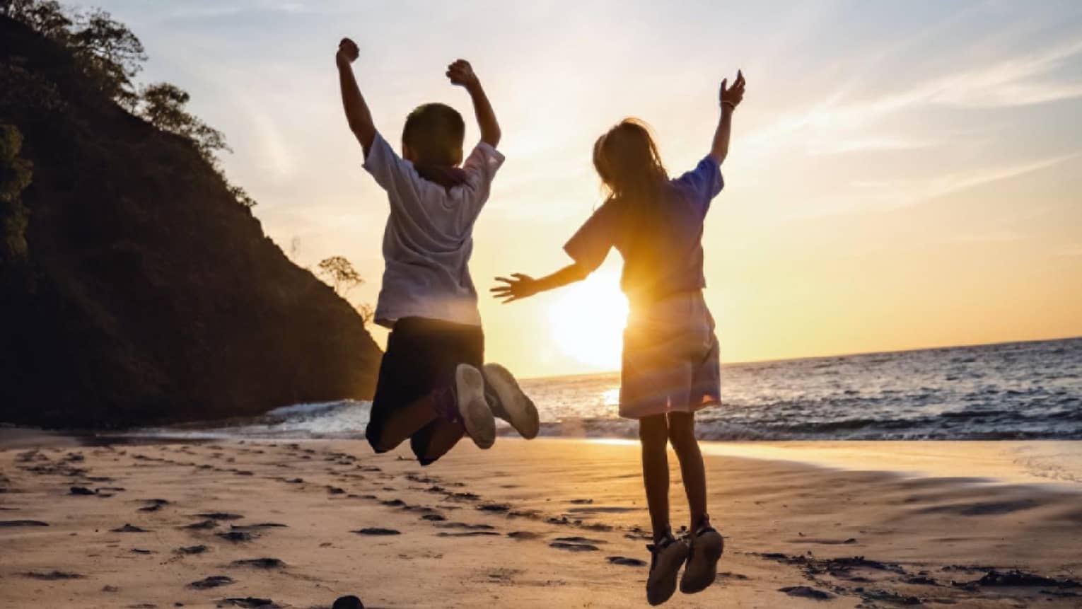 Two children jumping on a beach with the sun behind them.