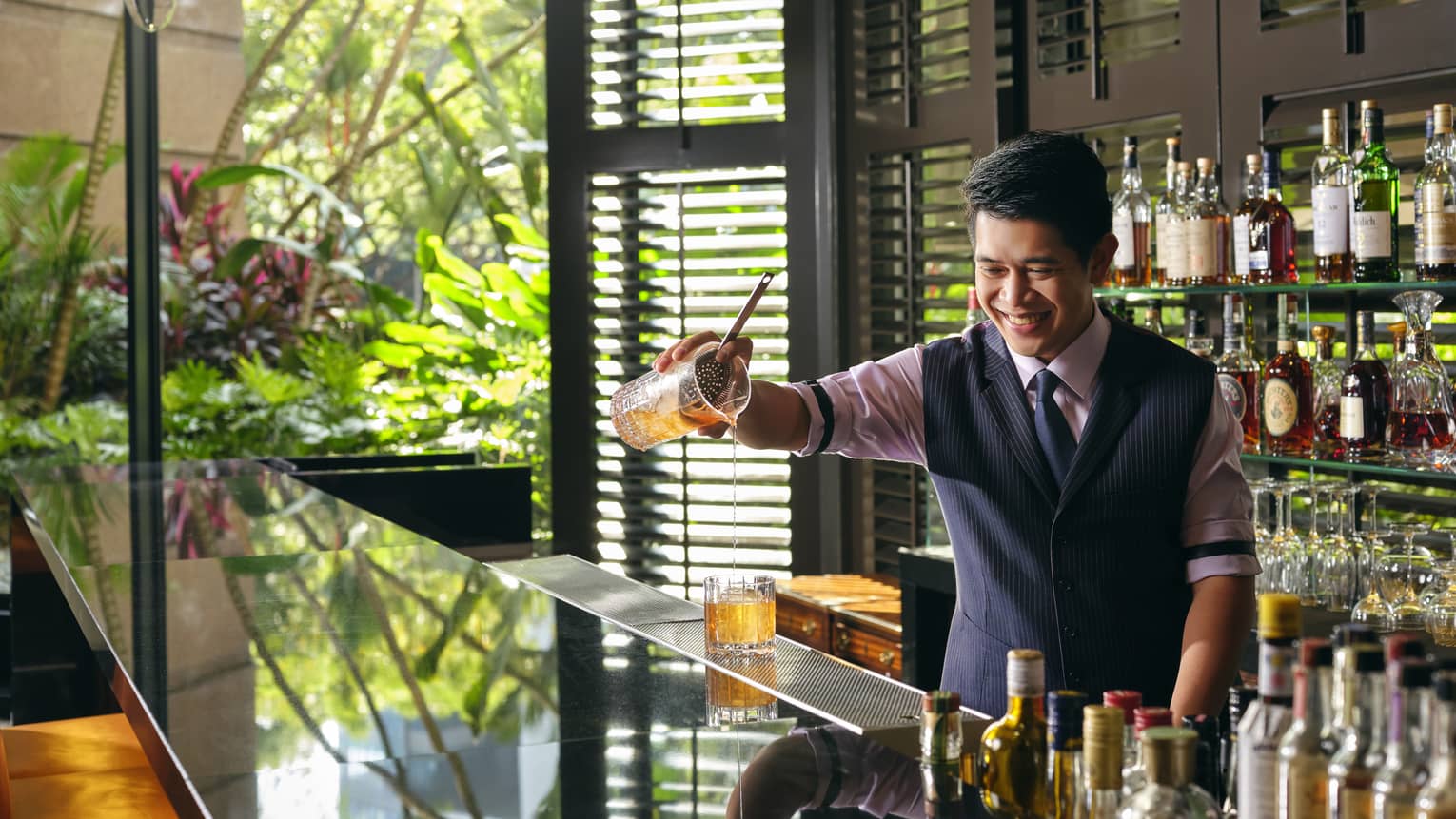A smiling bartender pours a Sidecar cocktail, a display of liquor bottles behind him, light streaming in from the windows to his right